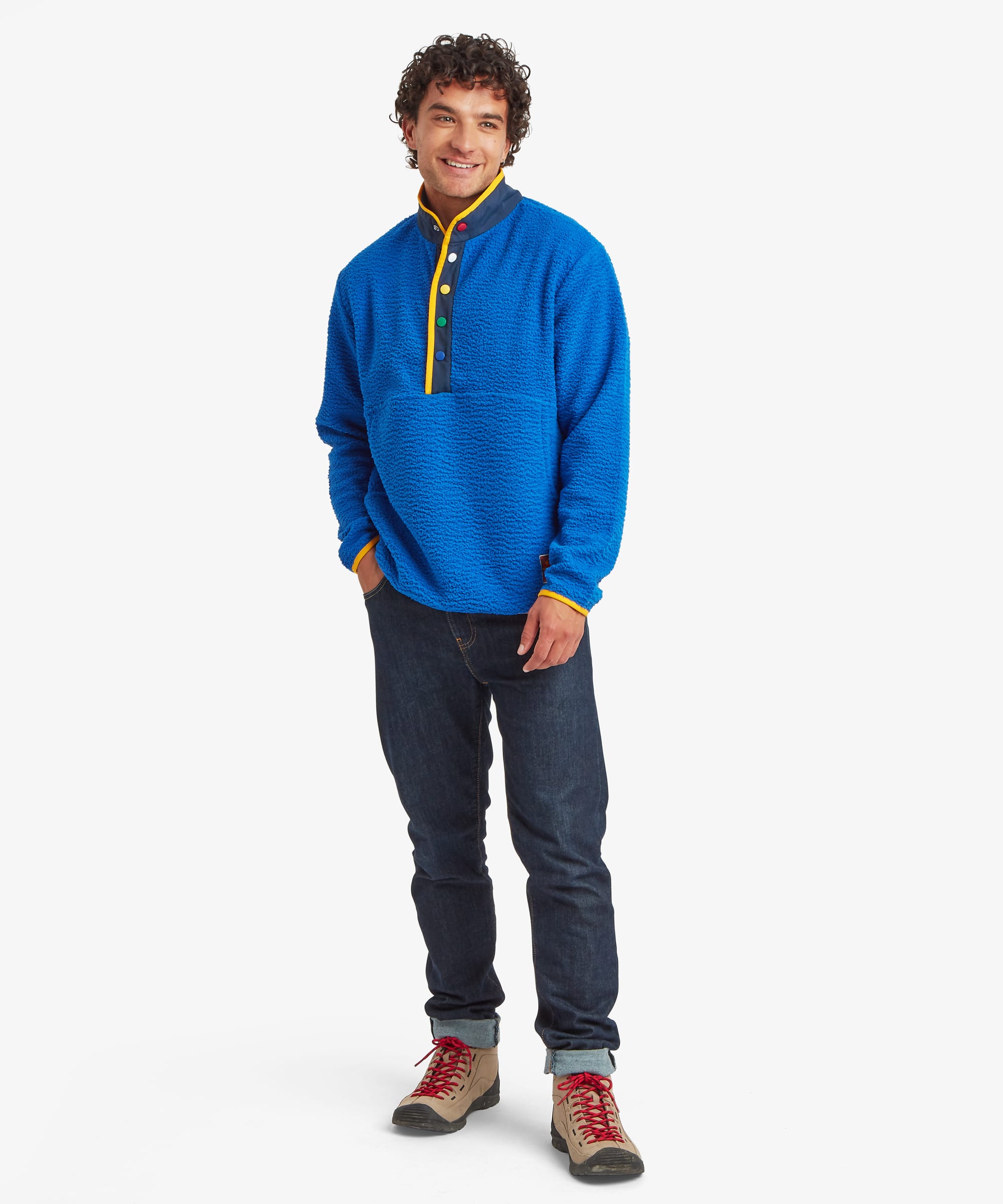 Full-length front view of a male model wearing the Sherpa Tarcho Snap Pullover in blue with dark blue jeans, showcasing the relaxed fit, textured fleece fabric, stand collar, colourful snap placket and hand pockets.