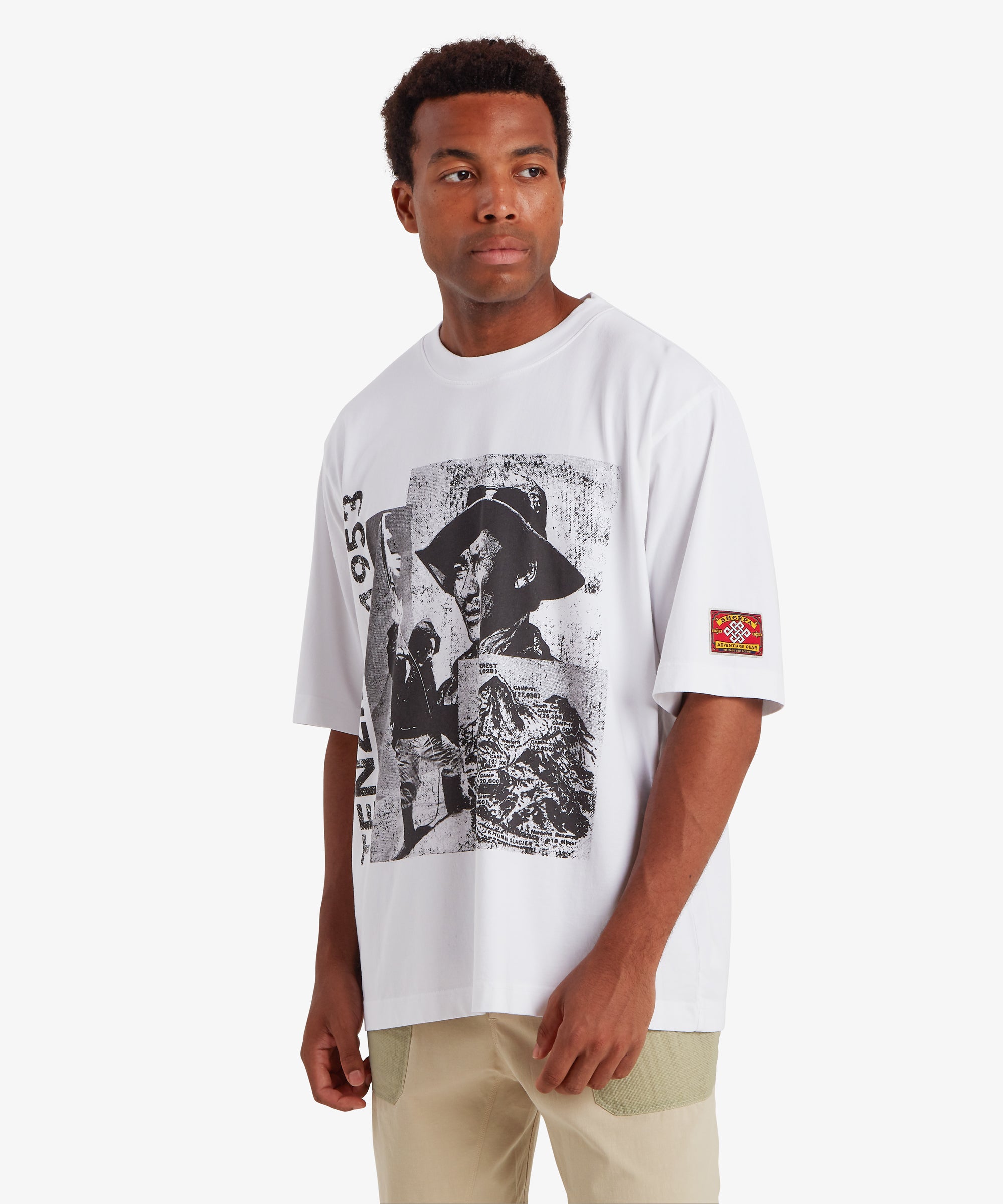 A man wearing a loose, relaxed fit white T-shirt. The T-shirt depicts a collage of three black and white picture of Tenzing Sherpa and Mount Everest. Next to the graphic, "Tenzing 1953" is written vertically on the left side of the T-shirt. On the the left sleeve is a red, mountaineering patch with a white endless knot in the centre surrounded by the words "Sherpa Adventure Gear". The outfit is completely with a pair of beige shorts with pale green pockets. The man is standing against a white background.