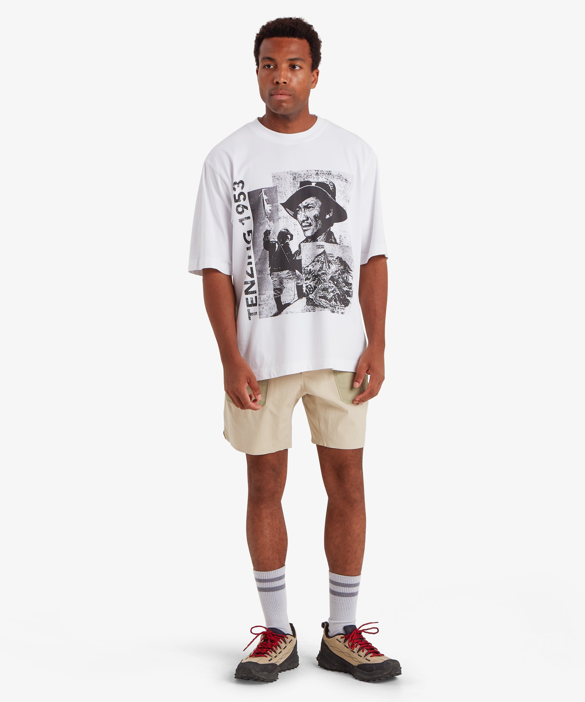 A man wearing a loose, relaxed fit white T-shirt. The T-shirt depicts a collage of three black and white picture of Tenzing Sherpa and Mount Everest. Next to the graphic, "Tenzing 1953" is written vertically on the left side of the T-shirt. The outfit is completely with a pair of beige shorts, white socks and beige walking shoes. The outfit is suitable for outdoor activities during summer. The man is standing against a white background with his hands down by his sides.
