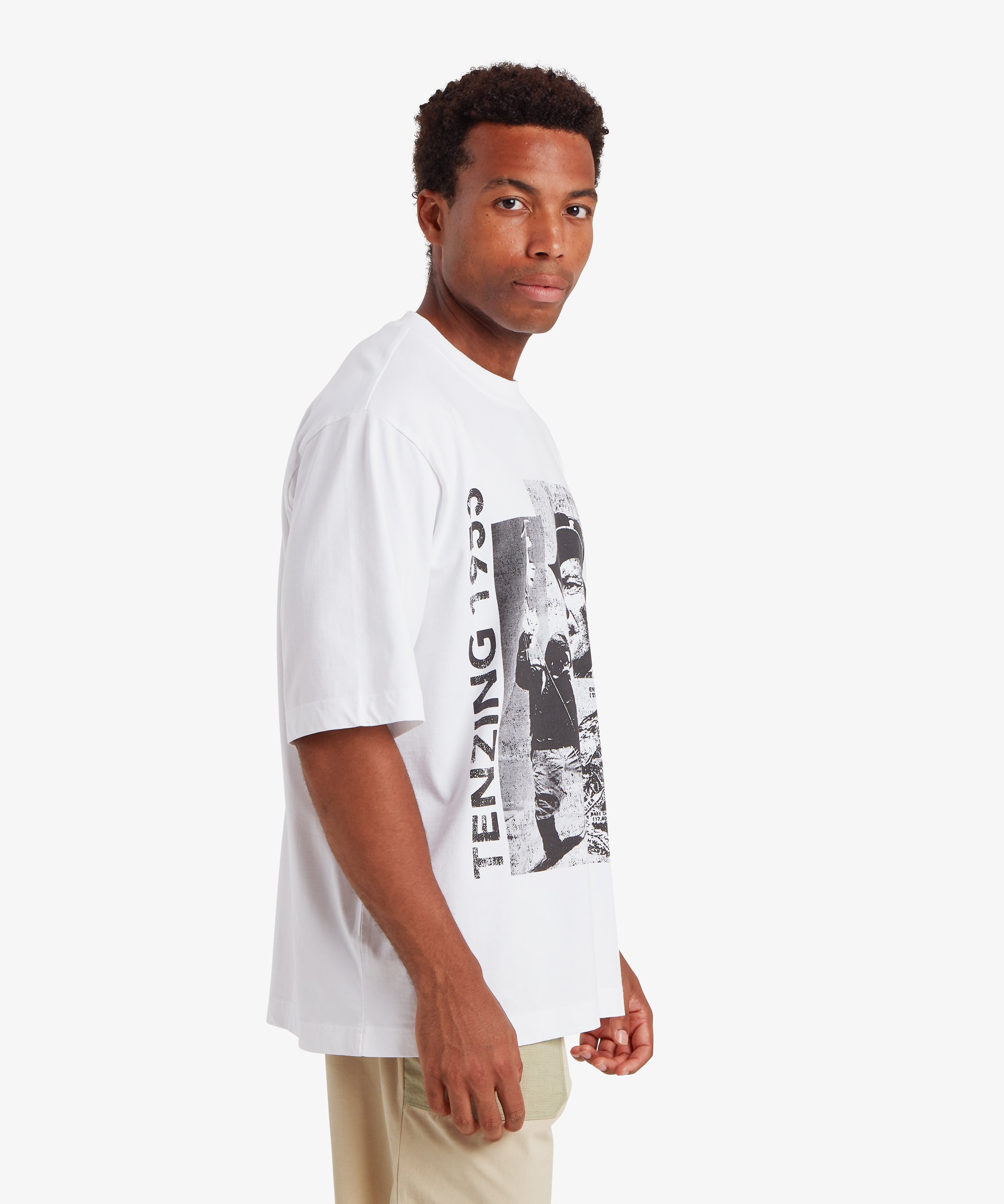 A man wearing a loose, relaxed fit white T-shirt. The T-shirt depicts a collage of three black and white picture of Tenzing Sherpa and Mount Everest. Next to the graphic, "Tenzing 1953" is written vertically on the left side of the T-shirt. A pair of beige shorts with pale green pockets compliment the T-shirt. The outfit is relaxed and suitable for everyday wear. The man is standing against a white background with his body turned to the left and his hands down by his sides.