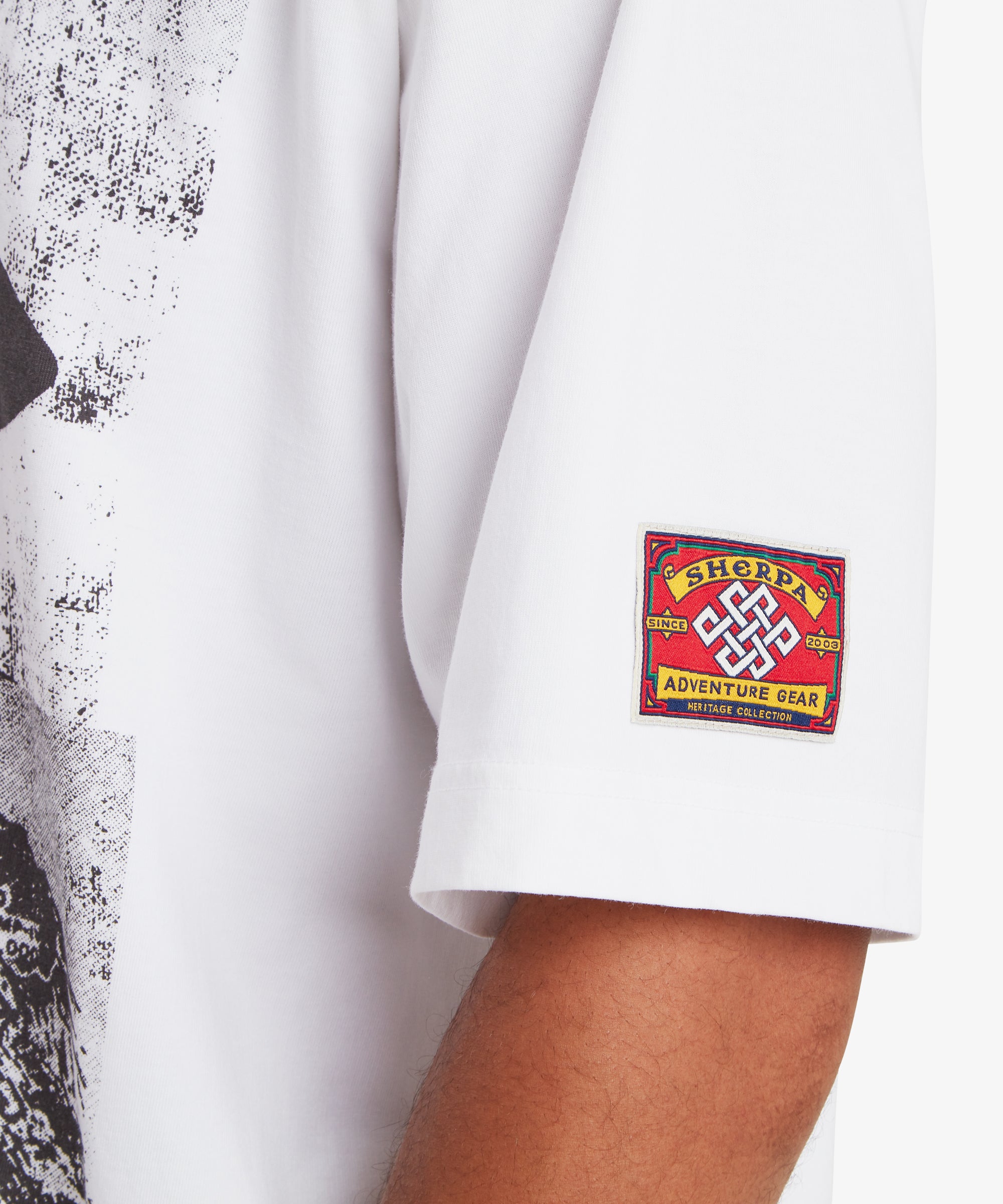 A close up of a sleeve of loose fit white T-shirt. Stitched on the sleeve of a T-shirt is red heritage mountaineering patch. At the centre of the patch is white Nepalese endless knot. Above and below the knot are the words "Sherpa Adventure Gear" written on a yellow background. Either side of the knot are the words "Since 2003" also on a yellow background. At the very bottom of the patch are the words "Heritage collection". The front of the T-shirt can also be seen with a graphic portrait of Tenzing Sherpa.