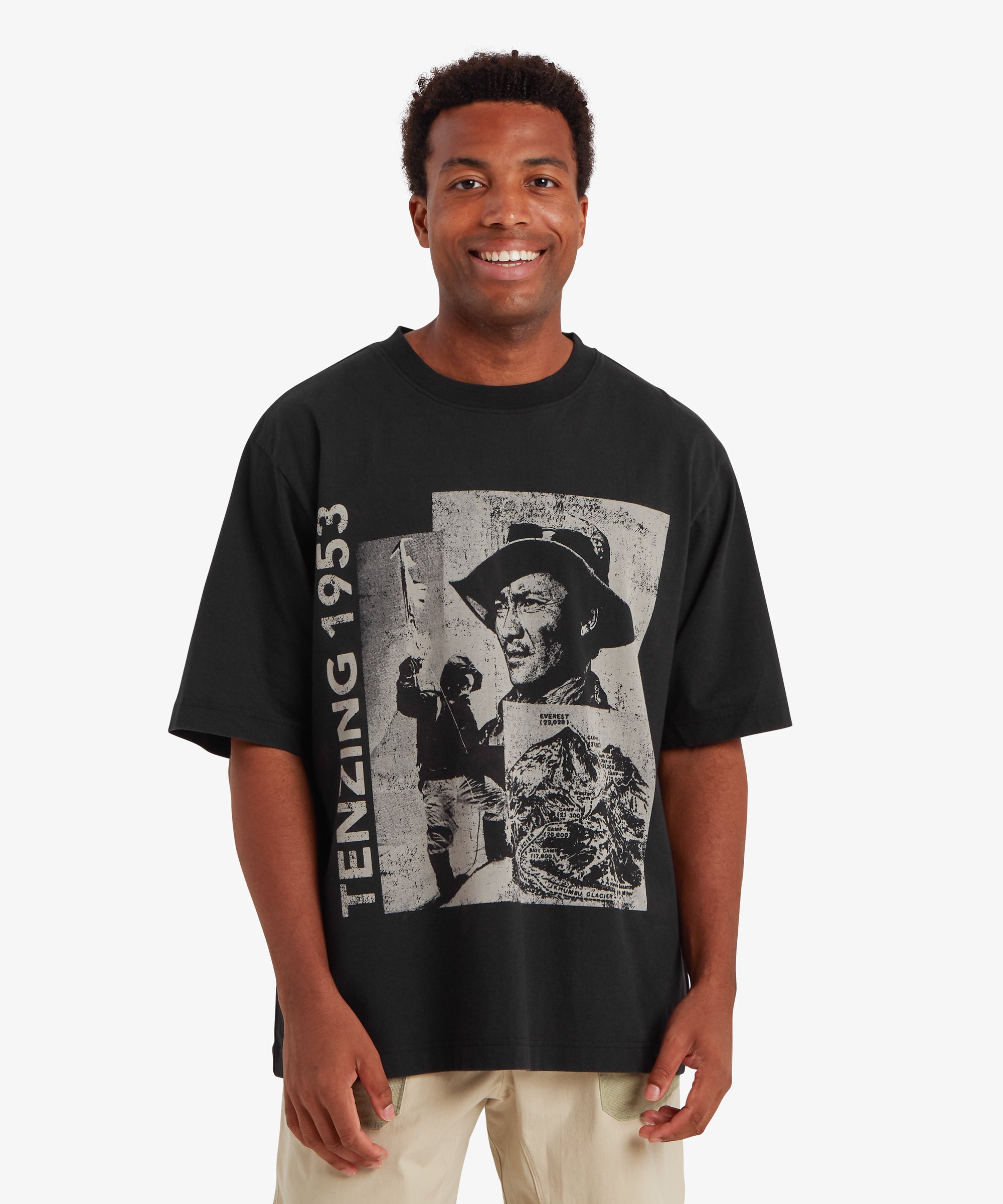 A man wearing a loose, relaxed fit black T-shirt. The T-shirt depicts a collage of three greyscale pictures of Tenzing Sherpa and Mount Everest. Next to the graphic, "Tenzing 1953" is written vertically on the left side of the T-shirt. On the the left sleeve is a red, mountaineering patch with a white endless knot in the centre surrounded by the words "Sherpa Adventure Gear". The outfit is completely with a pair of beige shorts with pale green pockets. The man is standing against a white background, smiling