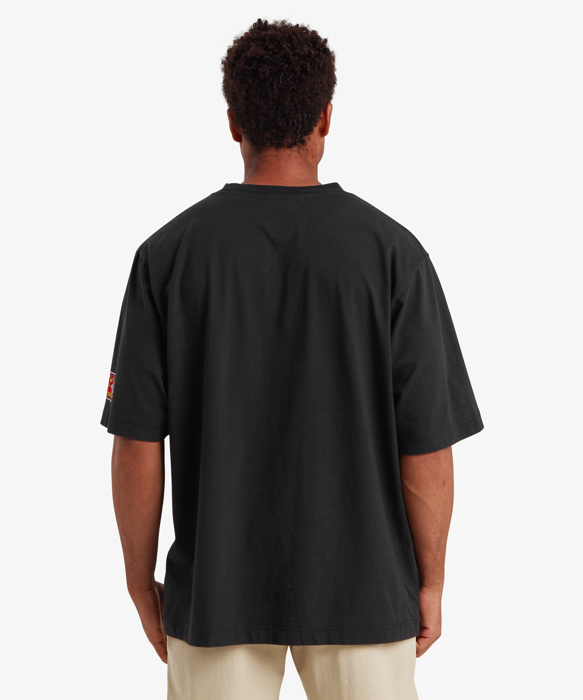 A back view of a man wearing relaxed fit Black T-shirt. A small red patch is stitched on the left sleeve of the T-shirt. The T-shirt is paired with beige shorts. The man is standing against a white background with his hands down by his sides.