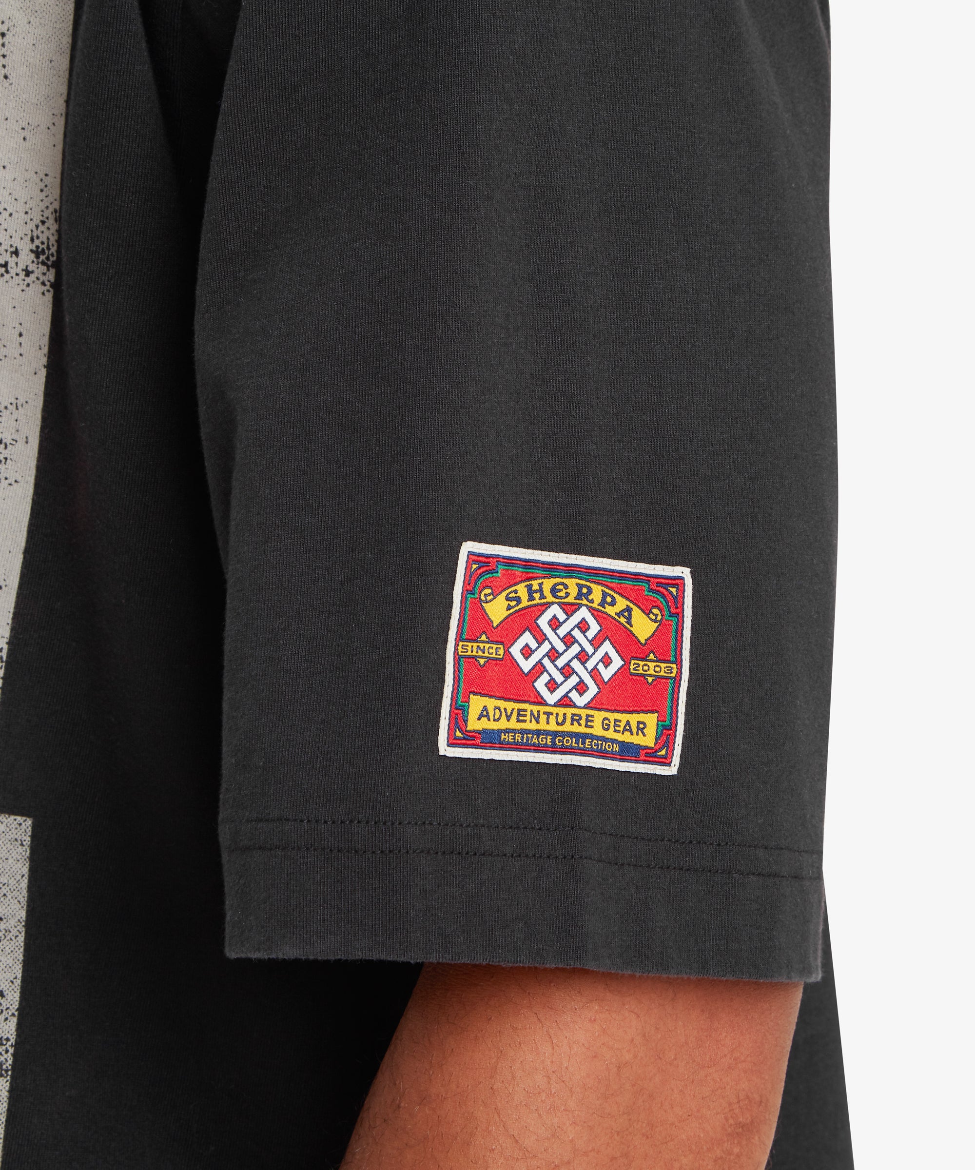 A close up of a sleeve of loose fit black T-shirt. Stitched on the sleeve of a T-shirt is red heritage mountaineering patch. At the centre of the patch is white Nepalese endless knot. Above and below the knot are the words "Sherpa Adventure Gear" written on a yellow background. Either side of the knot are the words "Since 2003" also on a yellow background. At the very bottom of the patch are the words "Heritage collection". A double line of stitching finishes off the hem of the sleeve.