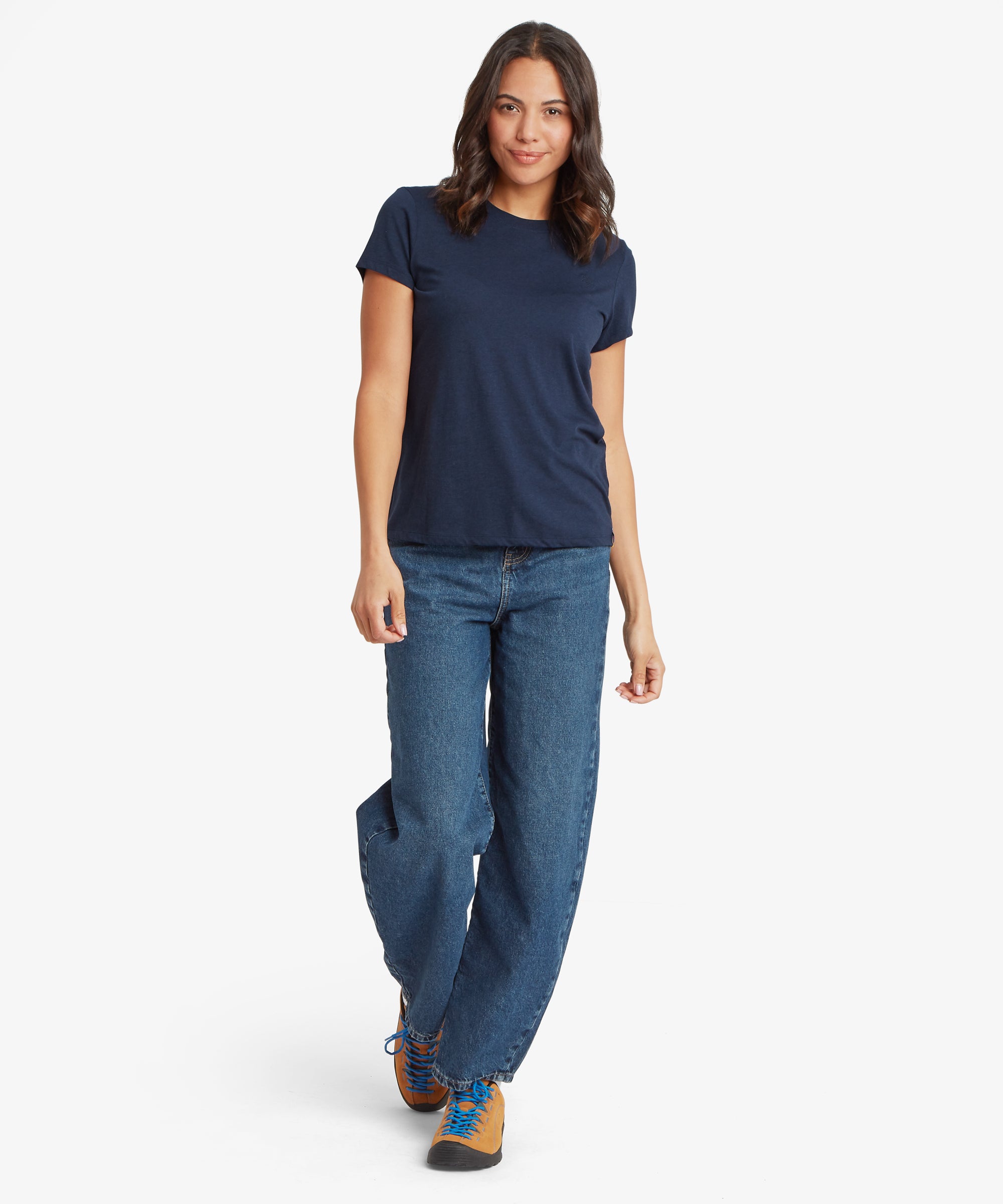 Full‑body front view of a person wearing a navy blue women’s short‑sleeve crew‑neck T‑shirt paired with relaxed‑fit mid‑wash denim jeans and tan hiking shoes with blue laces. The T‑shirt has a smooth, lightweight fabric with a classic hip‑length cut, shown clearly from the front as the person walks forward. The image highlights the casual everyday fit, clean neckline, and overall silhouette of the navy tee.