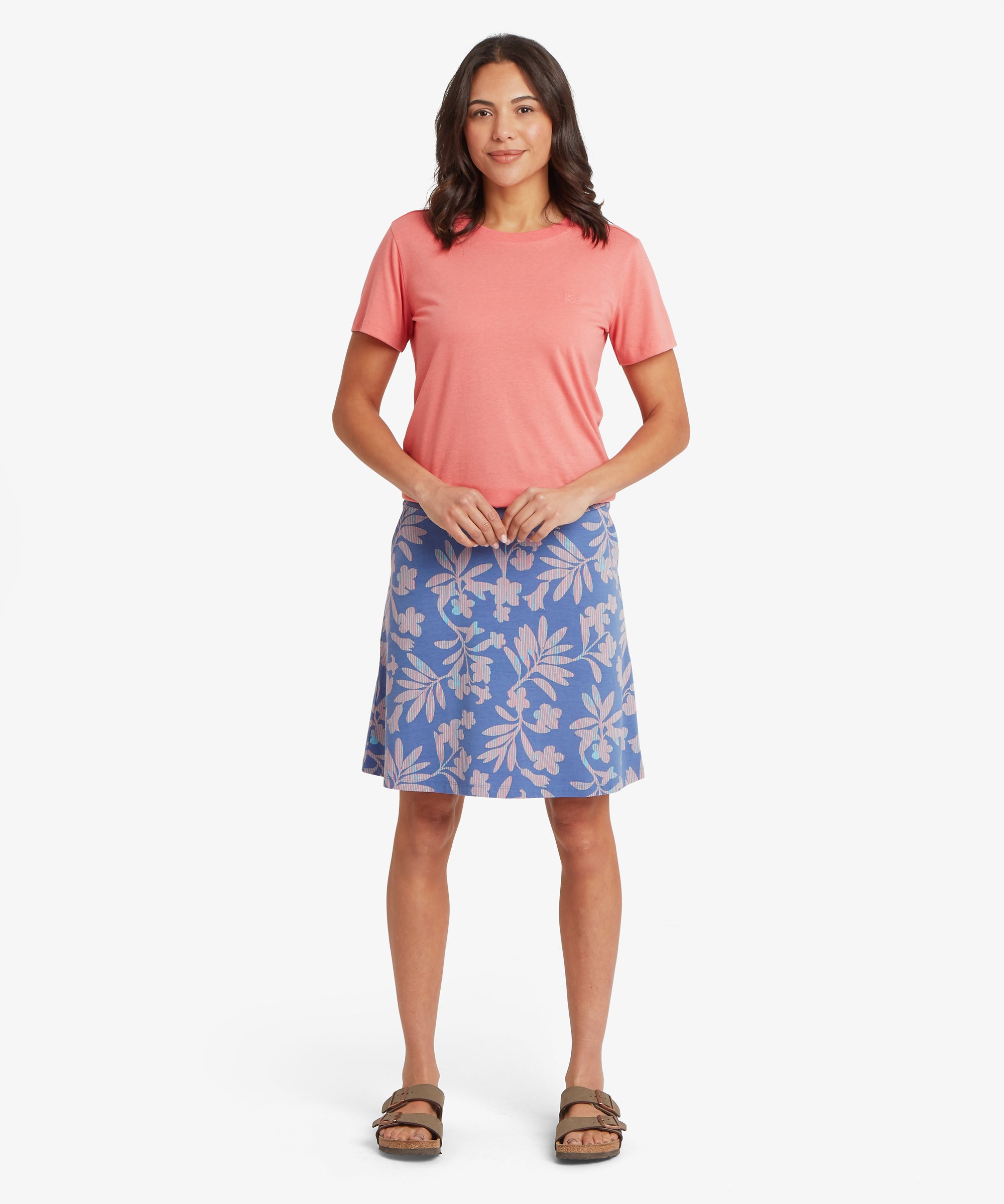 Front view of a coral pink short‑sleeve crew‑neck tee paired with a blue A‑line skirt featuring a large botanical leaf print, showing the relaxed drape, straight hemline, and lightweight jersey fabric styled with neutral sandals for an easy warm‑weather everyday outfit.