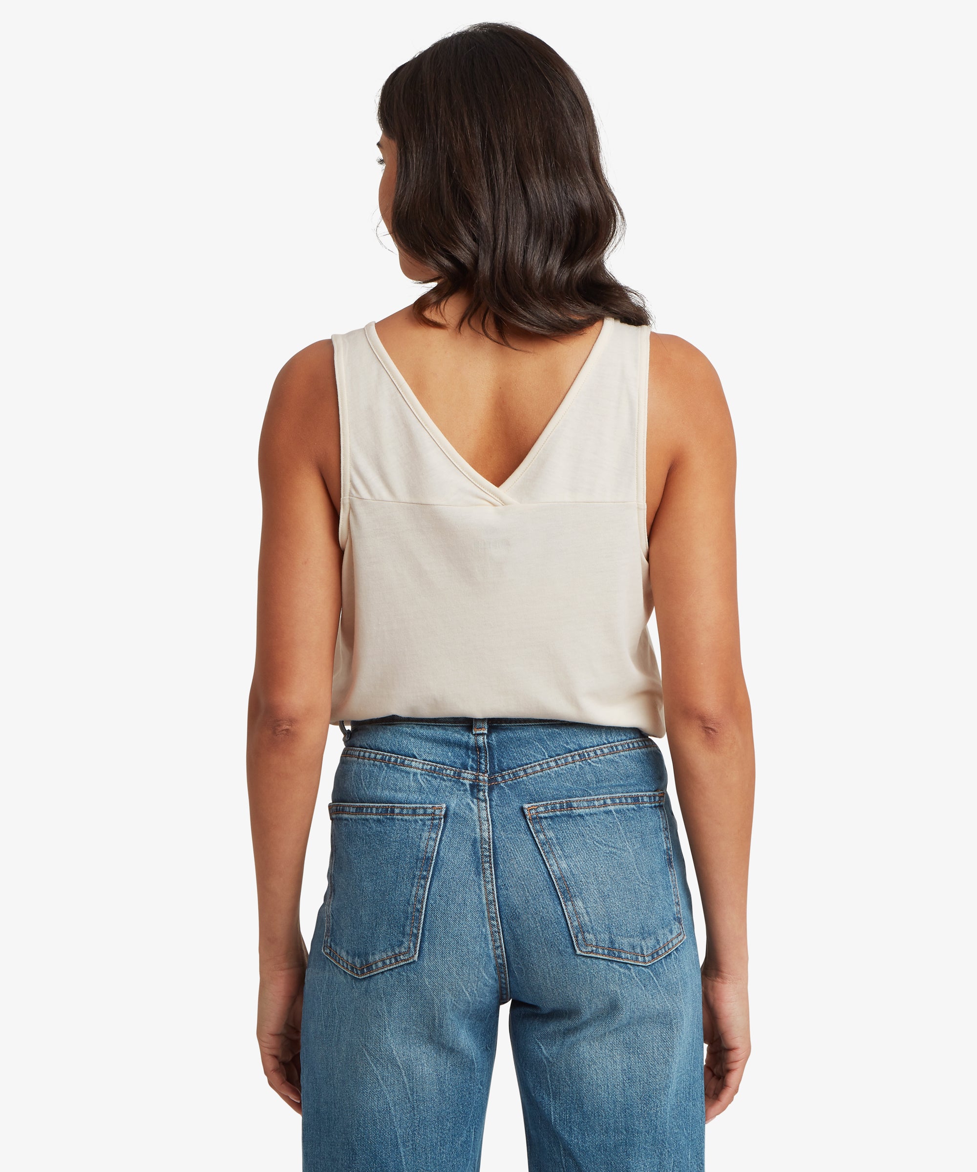 Back view of a cream women’s V‑neck tank top paired with mid‑wash denim jeans. The tank features a deep back V‑neckline, a clean back panel, and a relaxed fit that falls naturally to the hip.