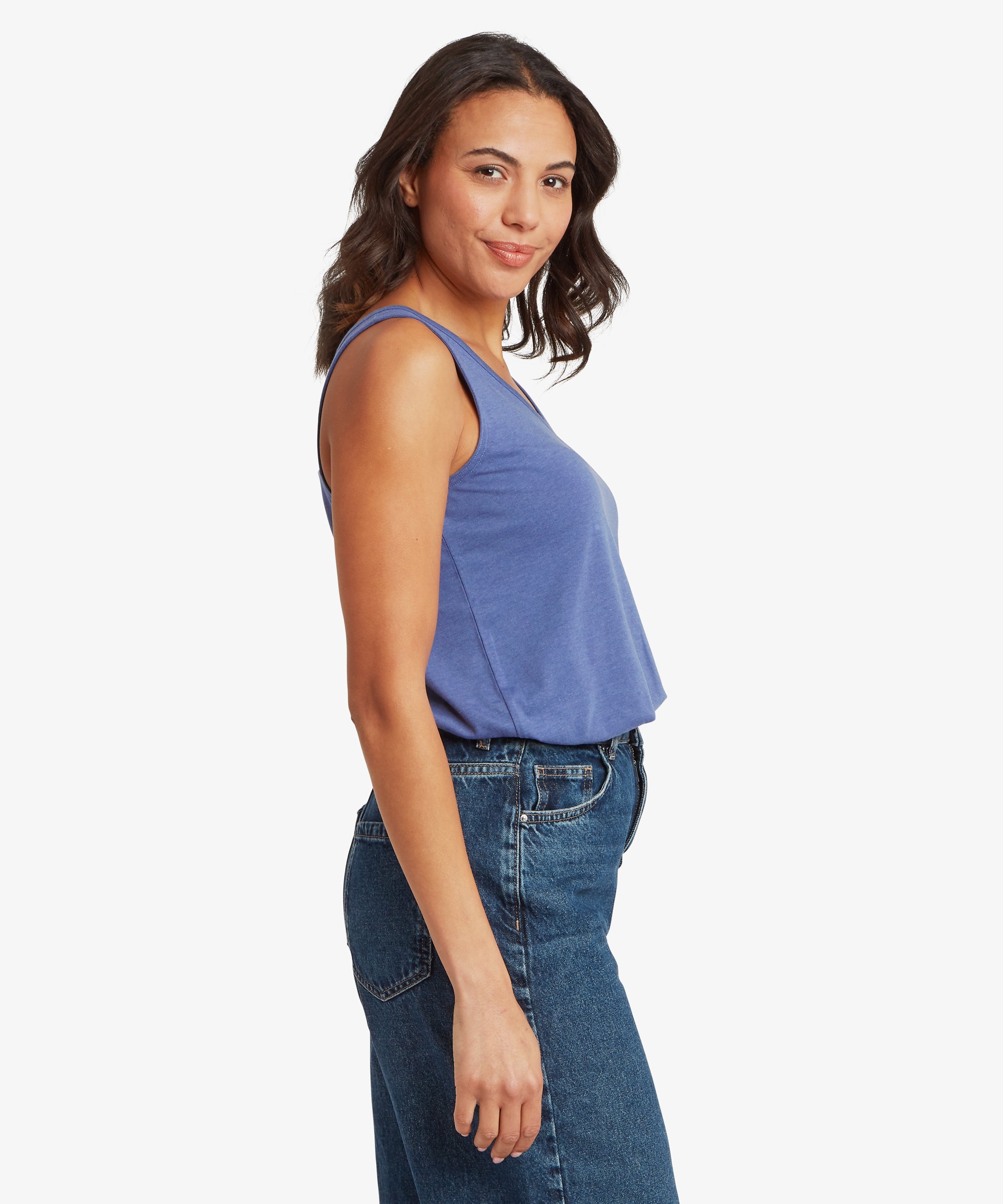Side view of a slate‑blue sleeveless V‑neck top paired with high‑waisted dark blue denim jeans, showing the smooth jersey fabric, relaxed drape, and softly curved neckline. One arm rests naturally by the hip, highlighting the comfortable warm‑weather styling ideal for casual everyday wear, travel, and lightweight summer layering.