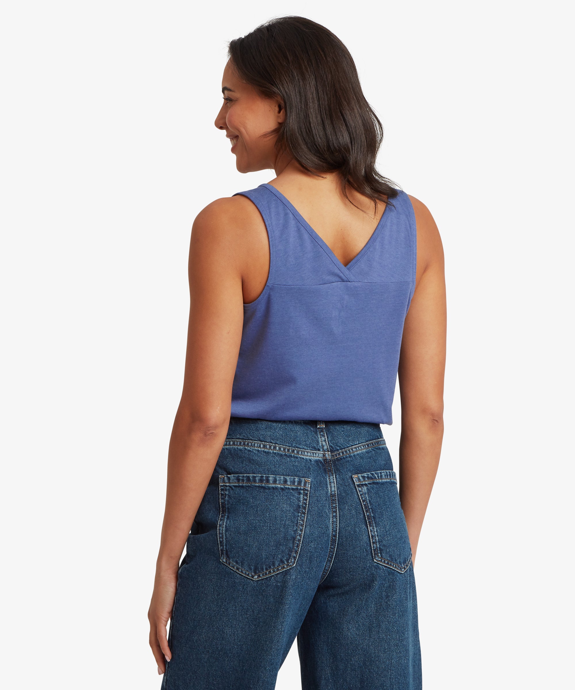 Back view of a sleeveless V‑neck top in a soft slate‑blue jersey fabric paired with dark blue denim jeans. The top features a gently curved V‑shaped back panel and a relaxed, easy drape. The hair falls naturally over the shoulders, and one arm hangs loosely by the side, highlighting the casual warm‑weather styling ideal for everyday wear, travel, and layered summer outfits.