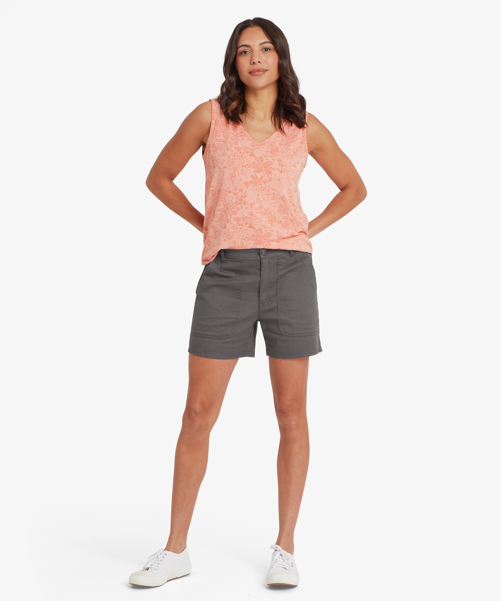 Full‑body view of a sleeveless V‑neck top in a soft peach patterned fabric paired with charcoal grey mid‑thigh shorts. The top has a relaxed drape and sits loosely over the waistband, while the shorts feature angled pockets and a straight hem. Both hands are placed on the hips, creating a clear front view of the outfit. White canvas trainers complete the look, highlighting a practical, comfortable warm‑weather style suitable for travel, walking, and casual everyday wear