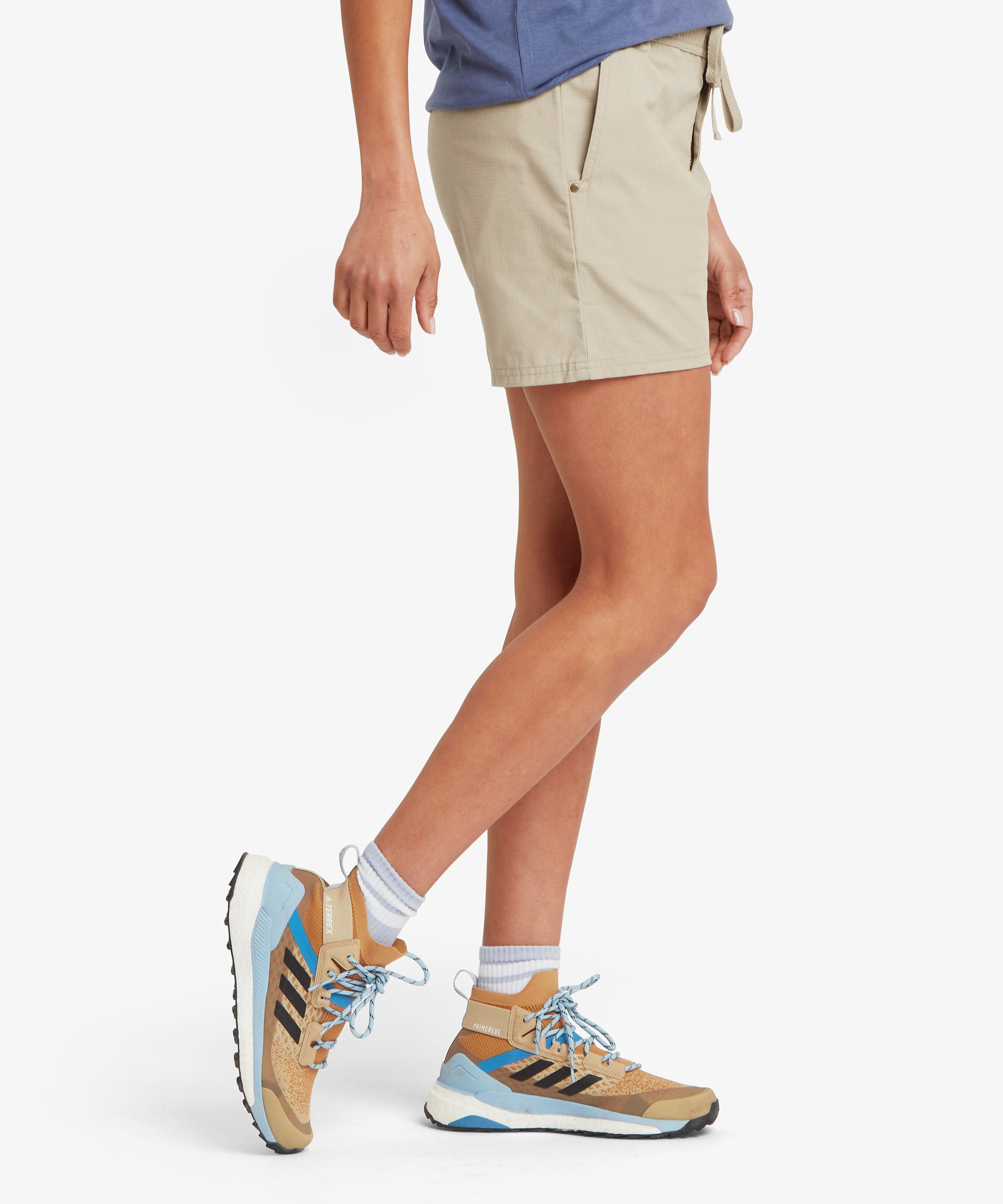 Side view of beige outdoor shorts paired with a slate‑blue T‑shirt and tan trail shoes with blue accents. One arm hangs naturally by the side while the forward leg position highlights the straight hemline, angled pocket design, and lightweight fabric. The overall outfit presents a functional, comfortable warm‑weather look suited for walking, hiking, and casual outdoor activity.