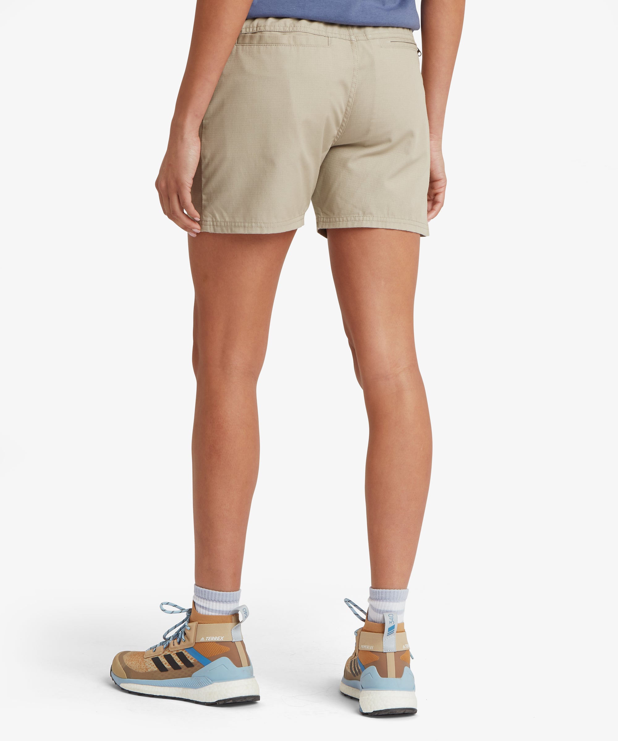 Rear view of beige outdoor shorts paired with a slate‑blue T‑shirt and tan trail shoes with blue detailing. The shorts feature a smooth elastic waistband, a clean back panel without visible pockets, and a straight mid‑thigh hem. The posture clearly shows the relaxed fit and lightweight woven fabric designed for warm‑weather activity, travel, and everyday comfort