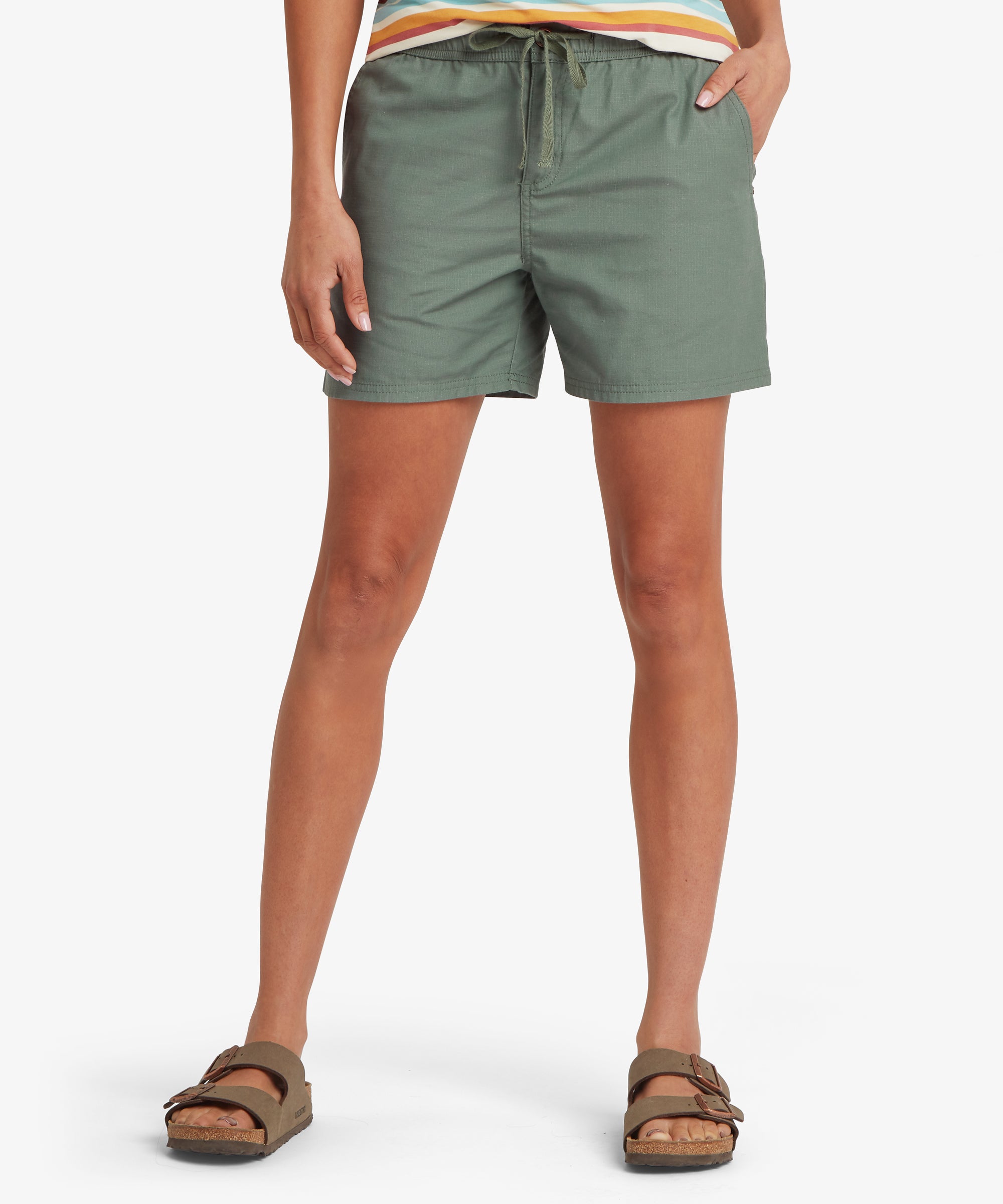 Front view of a light sage green outdoor skort showing the elastic drawstring waistband, angled seam detailing, and lightweight woven fabric. Paired with sandals, the skort’s relaxed fit and above‑knee length highlight a practical and comfortable warm‑weather style suited for everyday wear and travel.