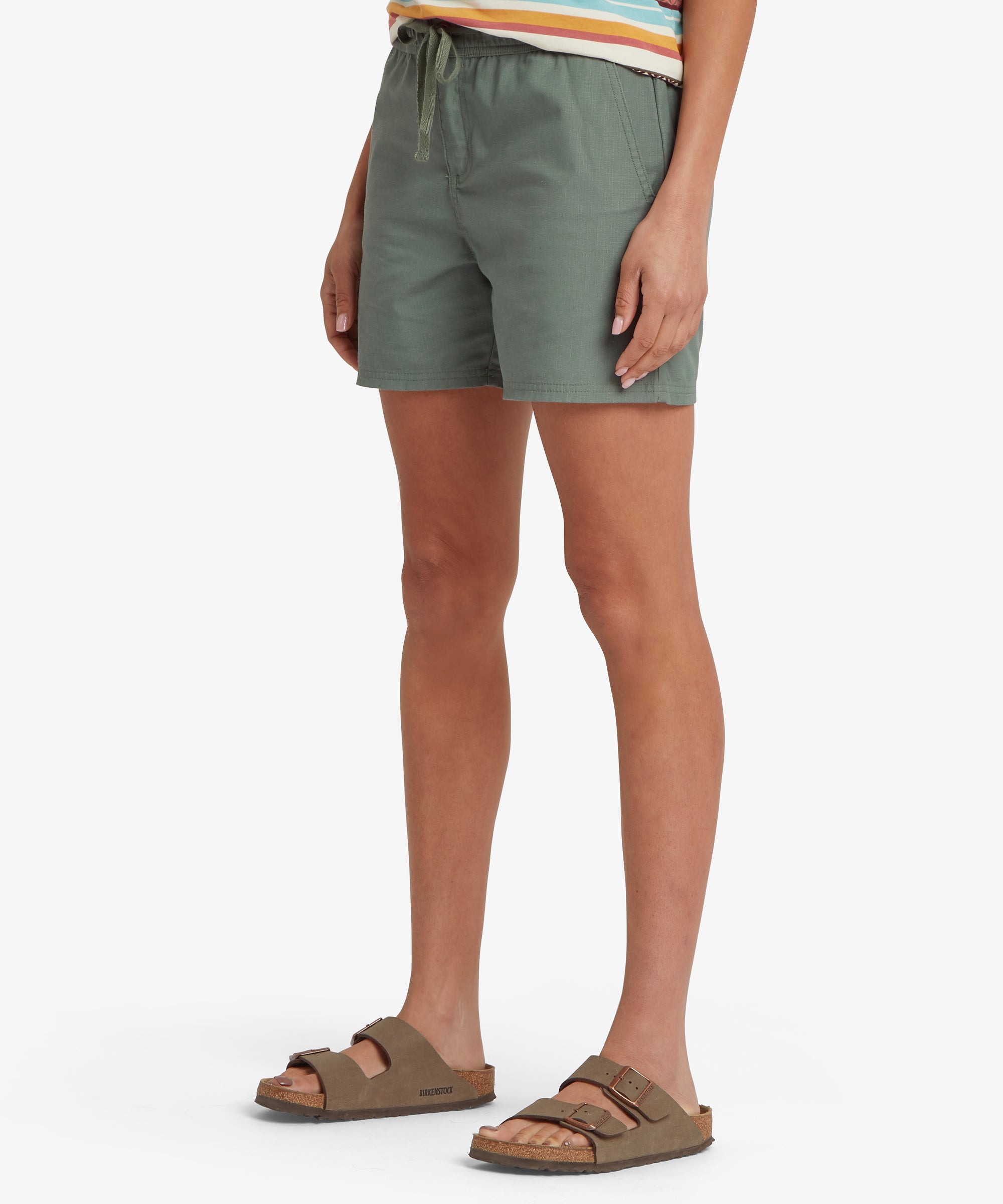Side view of a sage green pull‑on skort worn with a patterned top, showing the elastic drawstring waistband, patch hand pockets, and lightweight fabric. The relaxed fit and above‑knee length create a versatile warm‑weather option for travel, walking, and everyday comfort