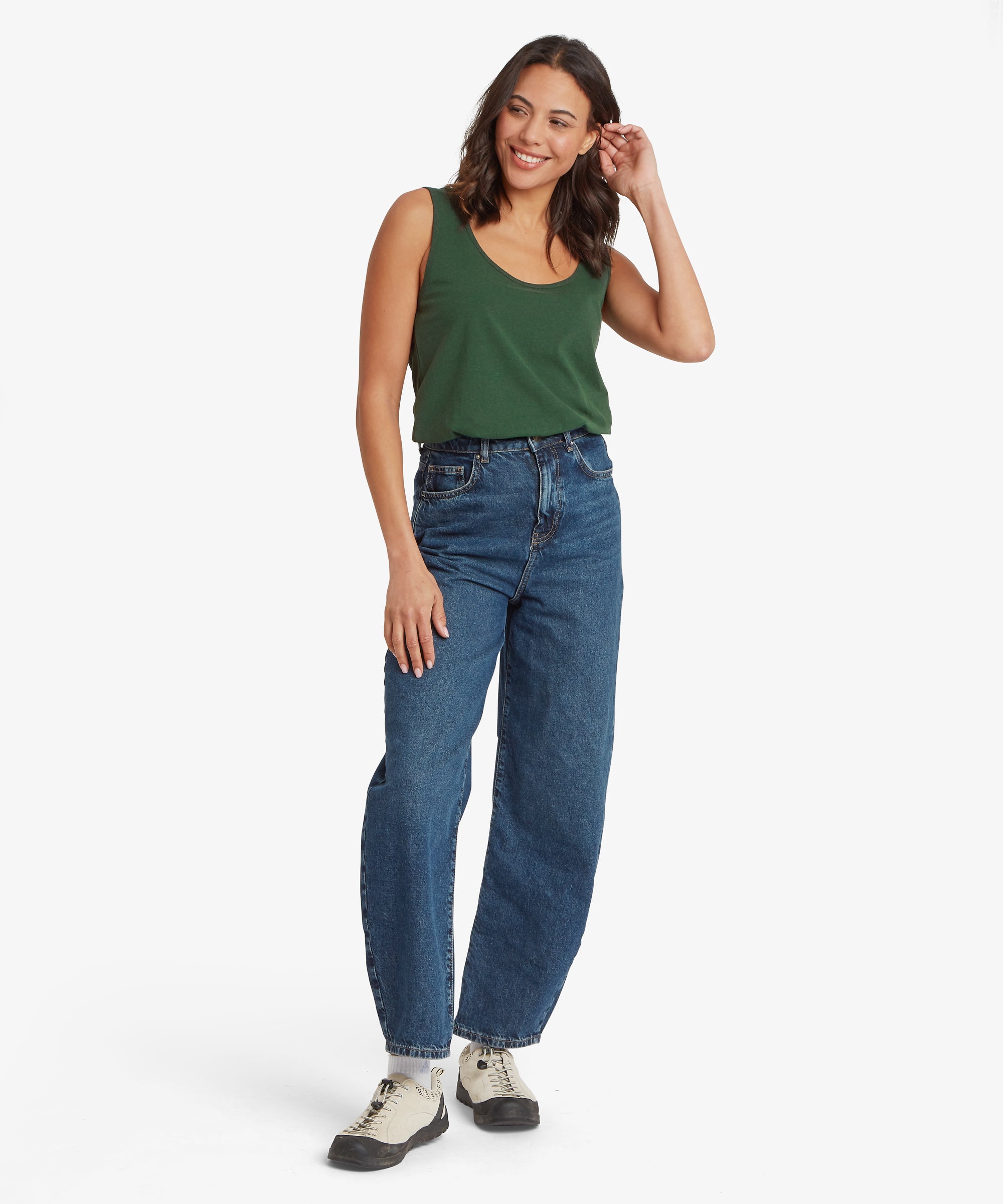 Full‑body view of an outfit featuring a dark green sleeveless scoop‑neck top paired with high‑waisted straight‑leg denim jeans. The top has a relaxed drape and tucks loosely at the waistband, while one arm hangs naturally and the other is raised lightly toward the hair. The jeans have a deep blue wash with visible seam detailing, and the look is completed with off‑white lace‑up boots with dark soles.