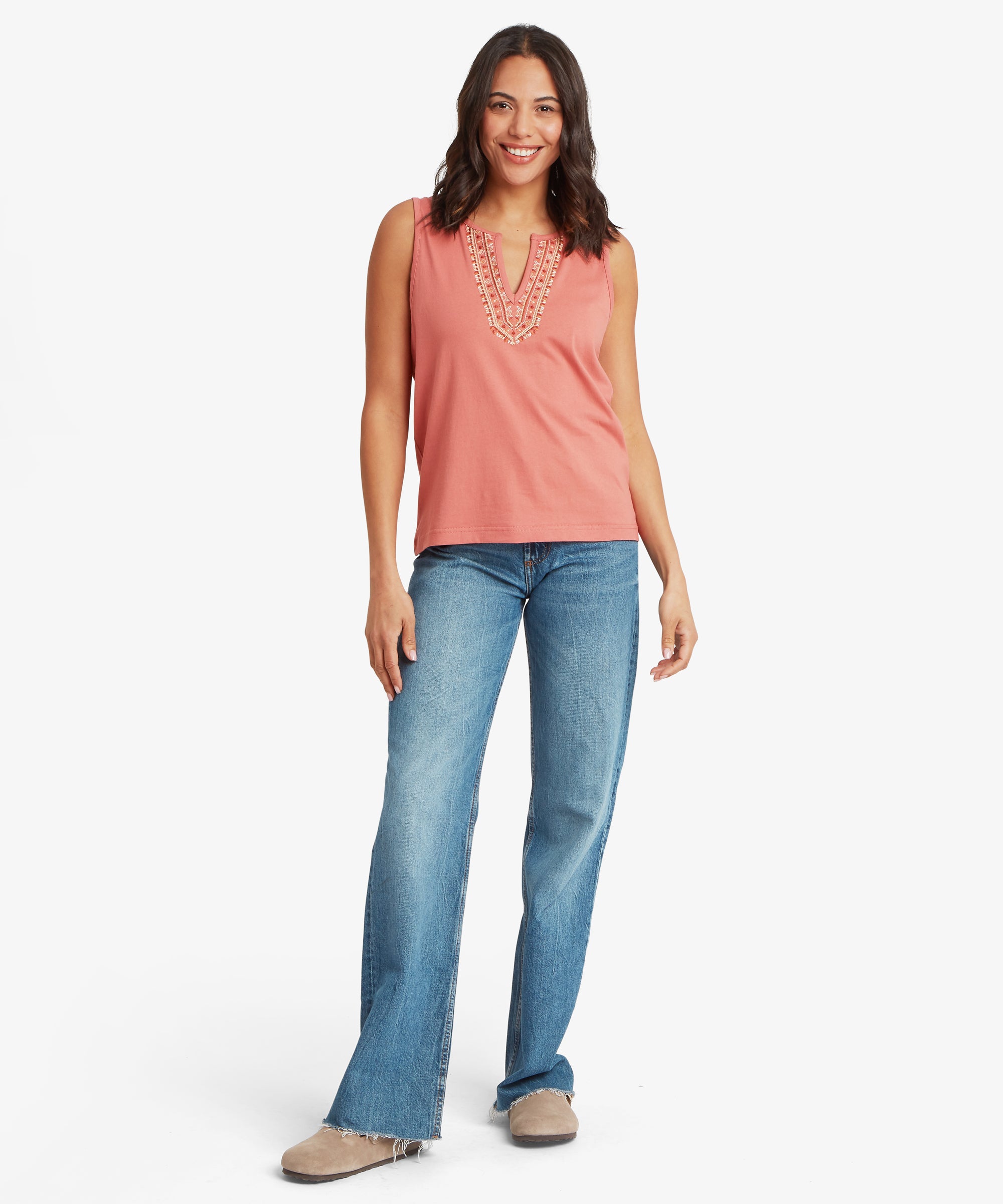 Full‑body view of a soft rose‑pink sleeveless top featuring an embroidered split‑neck detail, paired with mid‑wash blue flared jeans. The top drapes naturally over the waistband, showcasing the intricate stitched pattern at the neckline. One arm hangs loosely while the other is relaxed near the side, and the outfit is completed with neutral slip‑on shoes.