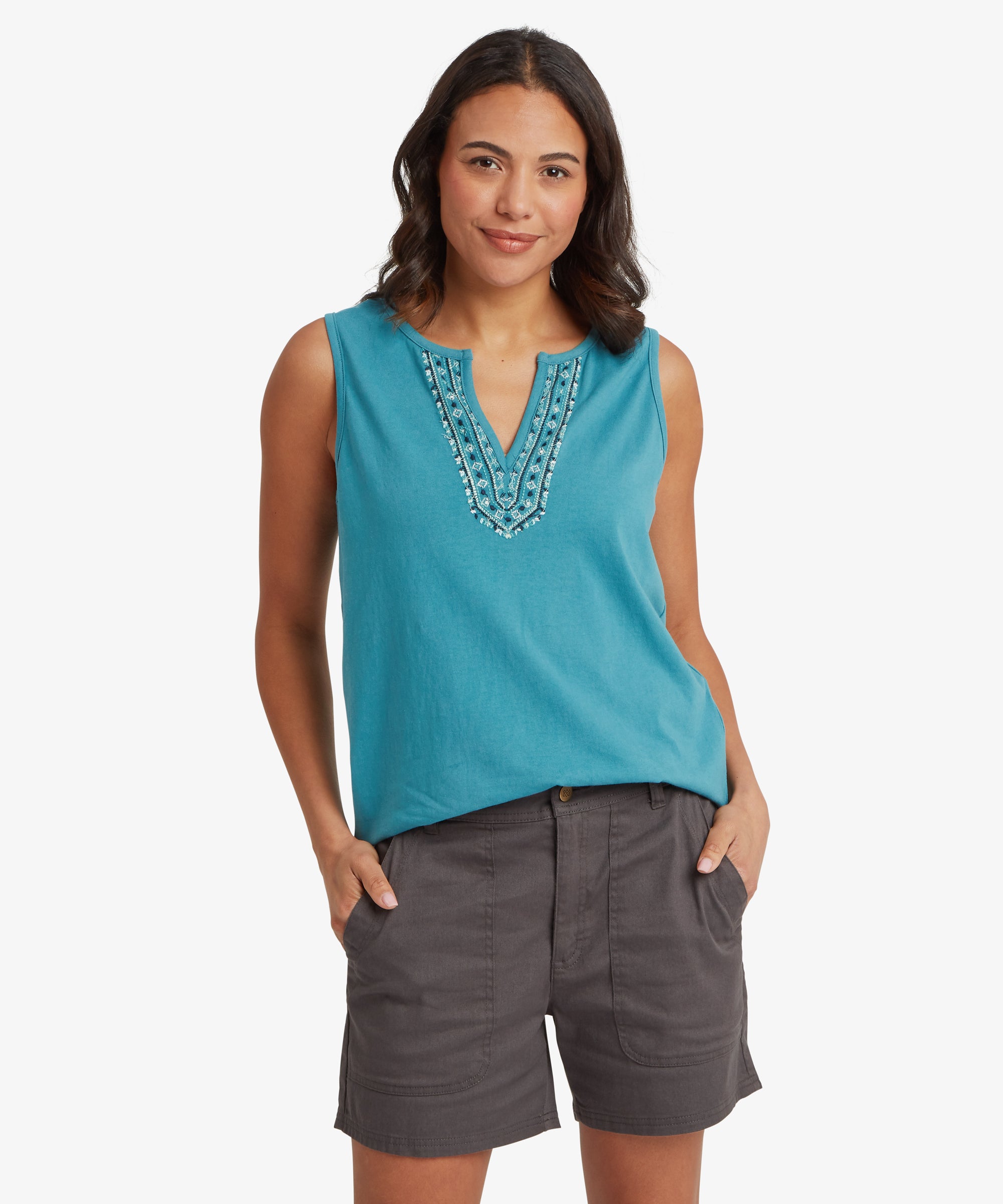 Front view of a turquoise sleeveless top featuring an embroidered split‑neck design with detailed layered stitching in coordinating blue and white thread. The top has a relaxed drape over charcoal grey mid‑thigh shorts, which include angled front pockets, reinforced seams, and a structured woven fabric. Both hands rest inside the pockets, and the outfit is paired with cork‑soled double‑strap sandals, highlighting a practical and comfortable warm‑weather look suitable for travel, walking, and casual everyday