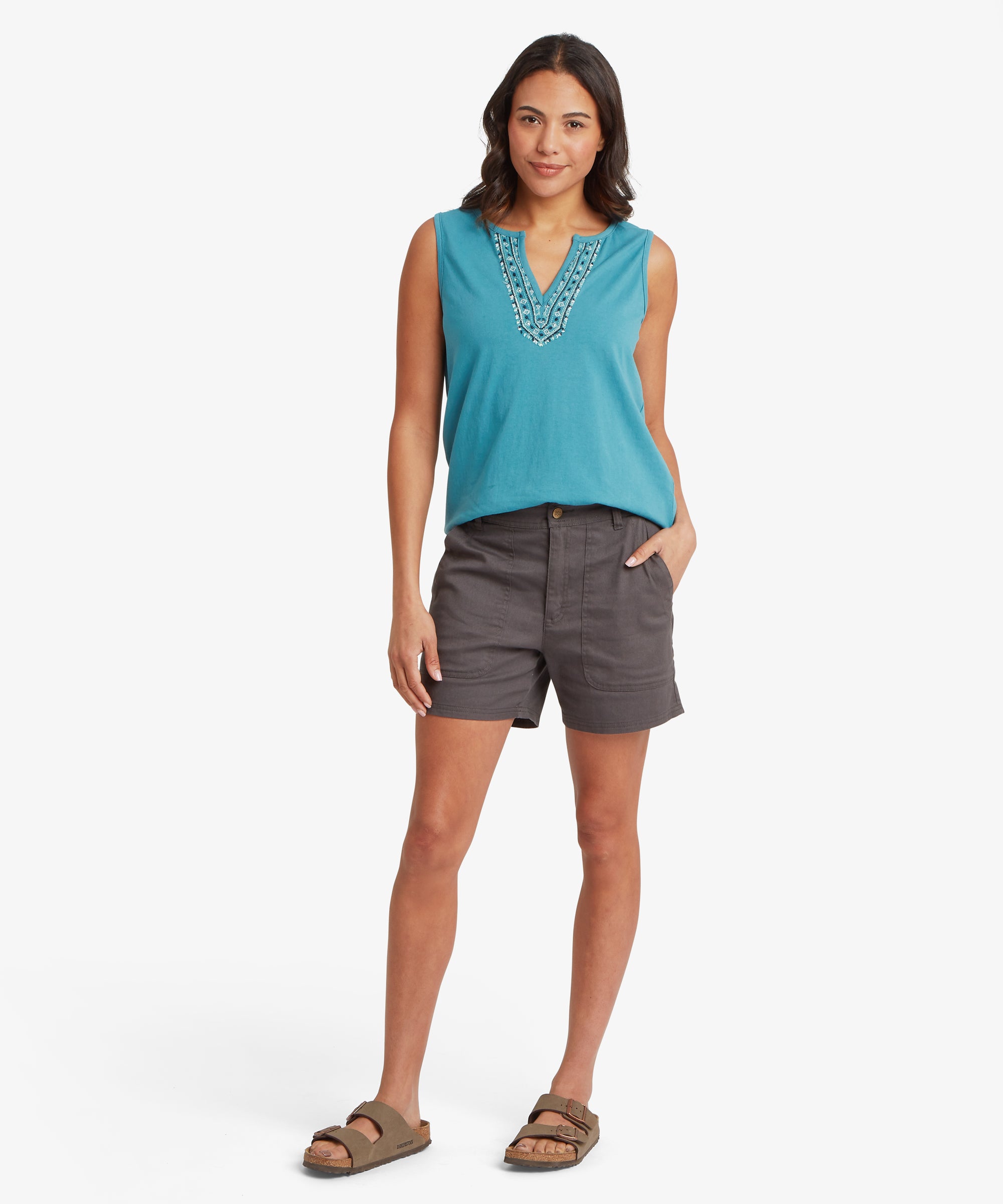 Full‑body view of a turquoise sleeveless top with an embroidered split‑neck design paired with charcoal grey mid‑thigh shorts. The top features intricate stitched detailing in coordinating blue tones, and the lightweight fabric drapes loosely over the waistband. One hand rests casually in a pocket while the other hangs naturally by the side.