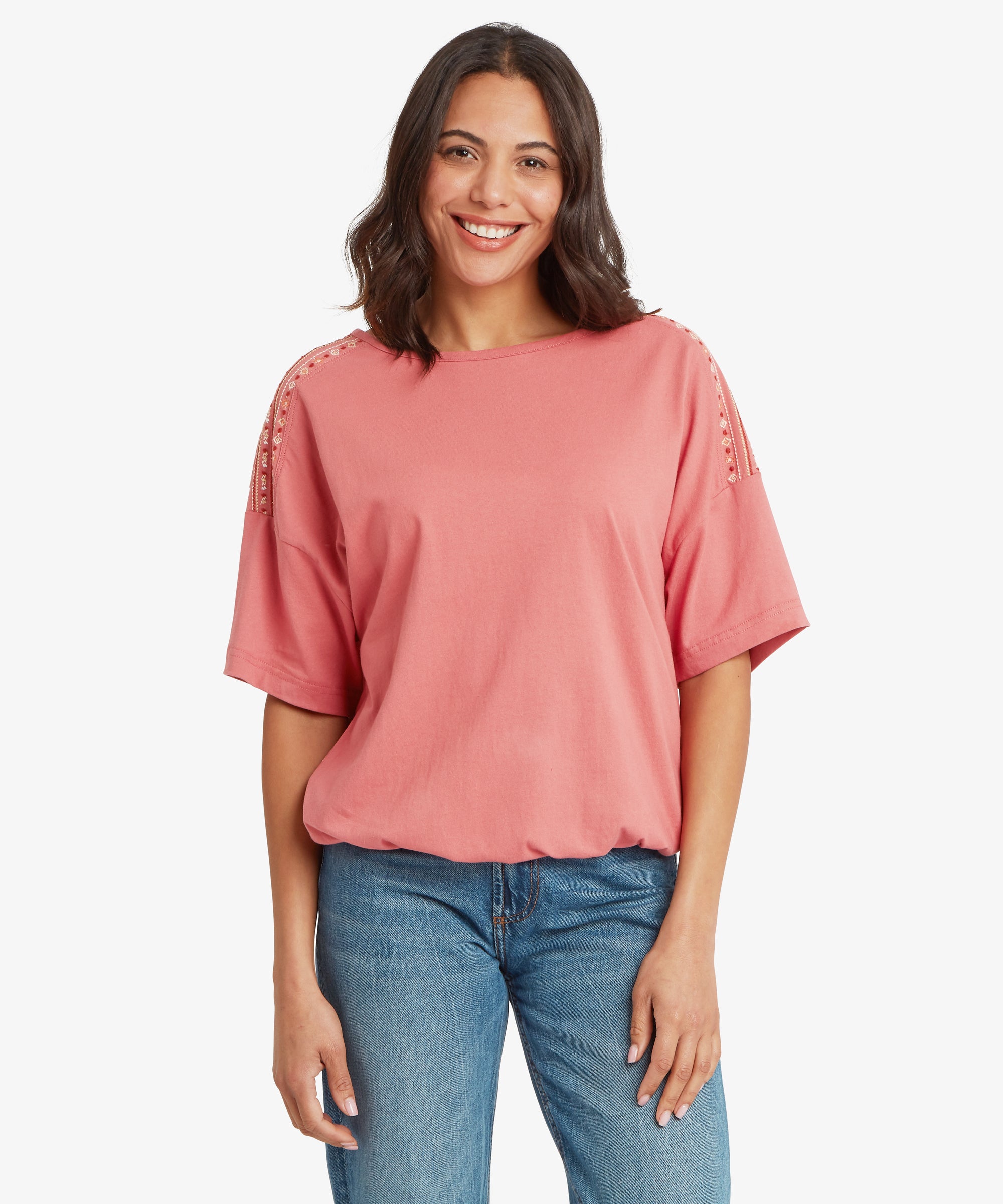 Front view of a rose‑pink short‑sleeve top with a relaxed, boxy fit and dropped shoulders. The top features decorative embroidered panel detailing along the upper sleeves and has a soft drape that sits comfortably at the hips. It is paired with mid‑wash blue denim jeans with a straight‑leg cut and visible seam stitching. Both arms rest naturally at the sides, highlighting a casual warm‑weather outfit suitable for everyday wear, travel, and easy layering