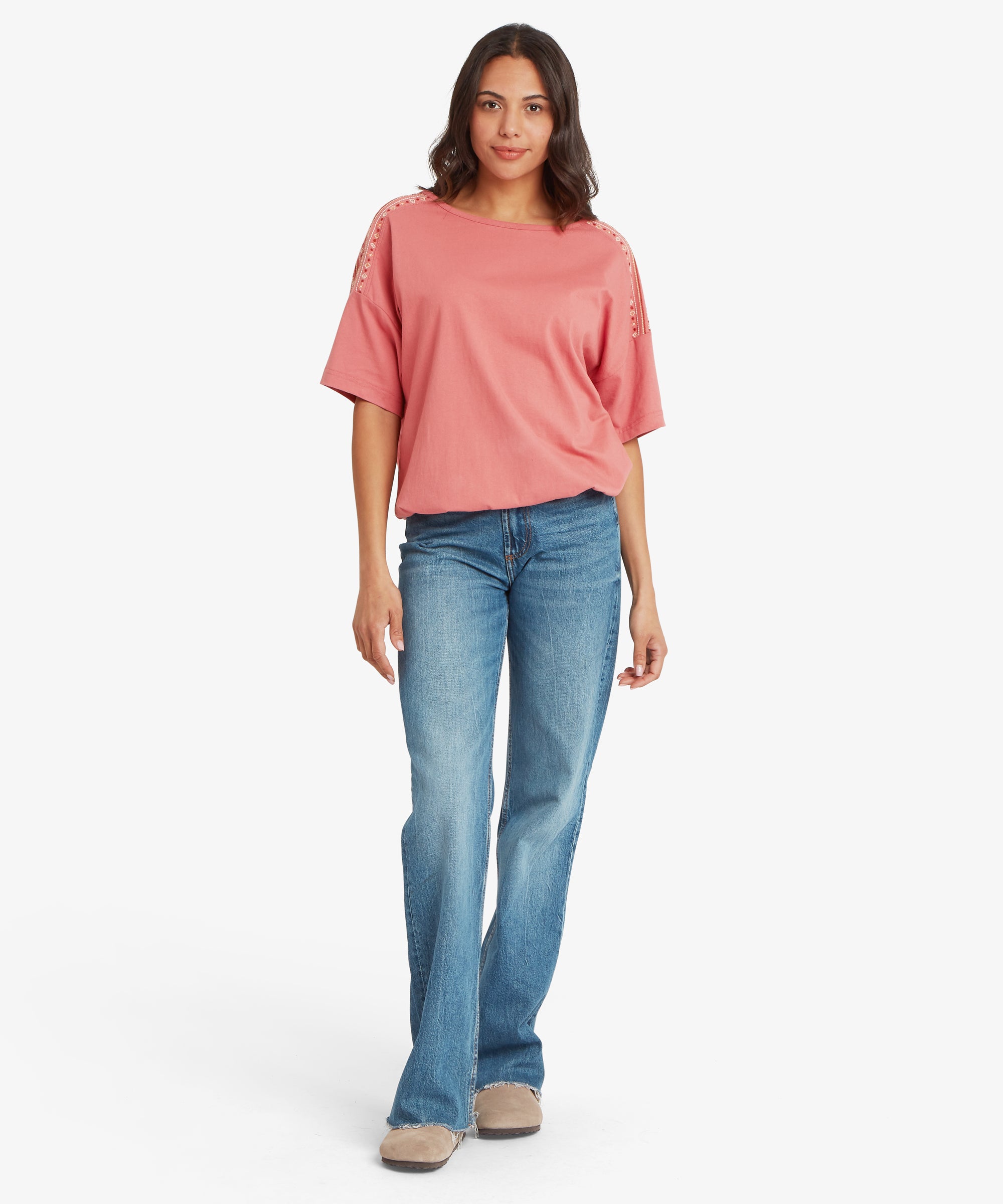 Full‑body view of a relaxed rose‑pink short‑sleeve top paired with mid‑wash blue flared jeans. The top features embroidered detailing along the shoulders and upper sleeves and has a loose, draped fit that sits comfortably at the hips. One arm hangs naturally by the side while the other rests slightly forward. The jeans have a classic straight‑through‑the‑thigh cut that widens below the knee, paired with neutral slip‑on shoes