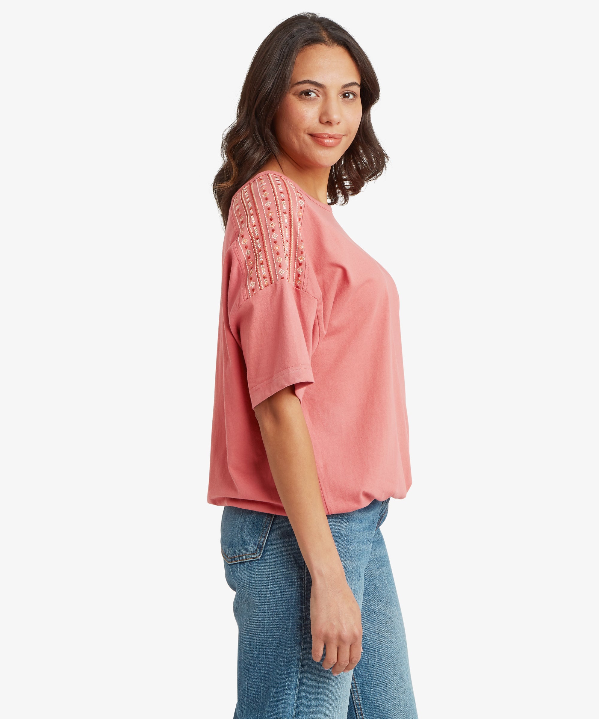 Side view of a loose‑fit short‑sleeve top in a soft rose‑pink colour, featuring decorative embroidered panels running along the shoulders and upper sleeves. The top has a relaxed drape and slightly curved hem, paired with mid‑wash blue jeans that include visible seam detailing and a classic straight‑leg fit. One arm hangs naturally by the side, highlighting a casual warm‑weather outfit suitable for everyday wear, layering, travel, and relaxed outdoor activities