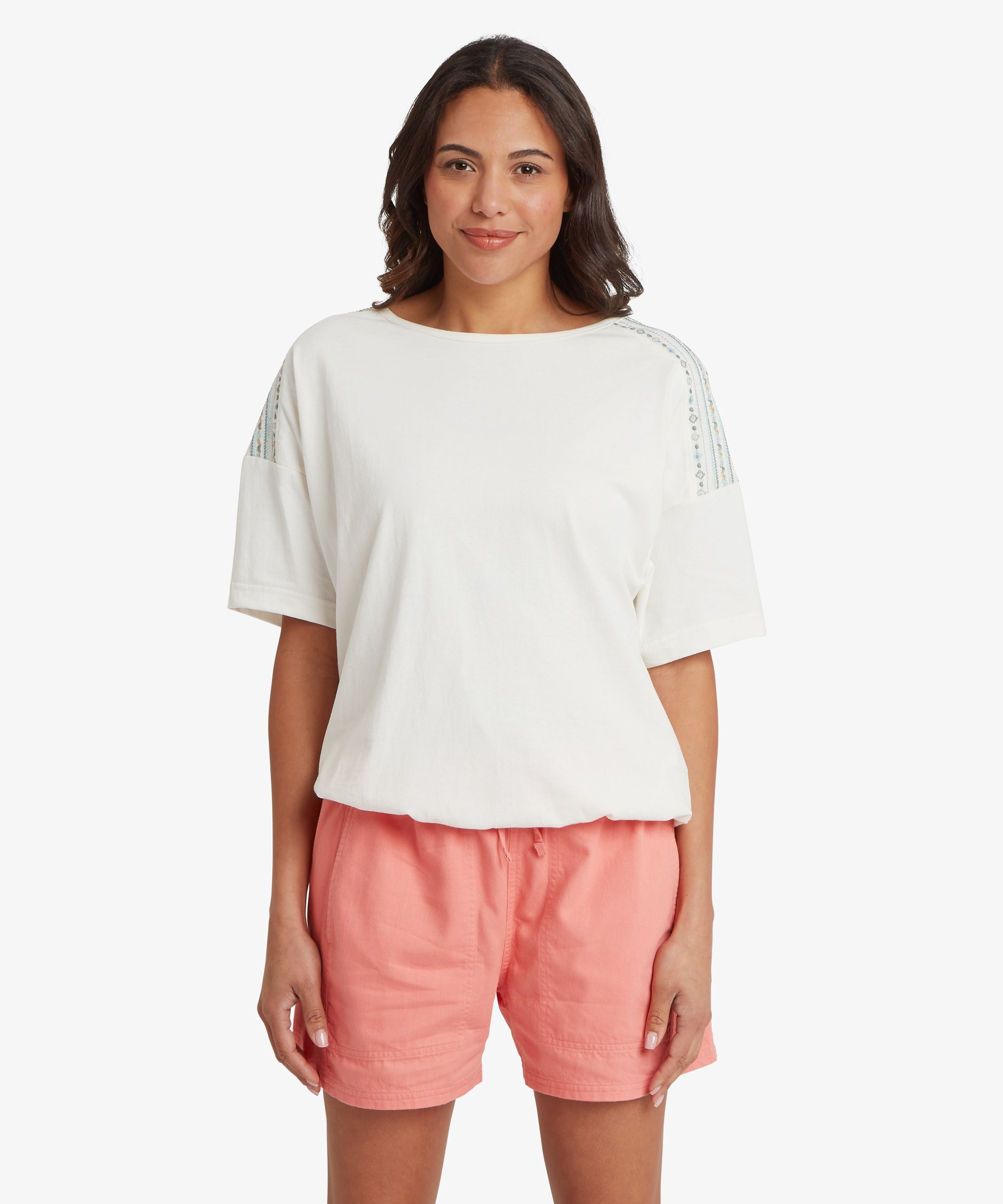 A person stands facing forward against a plain white background, wearing a loose white short‑sleeve top with vertical patterned stripes running along each shoulder. The top has a relaxed drape and a round neckline. They pair it with coral shorts featuring an elastic waistband and a comfortable, easy fit.