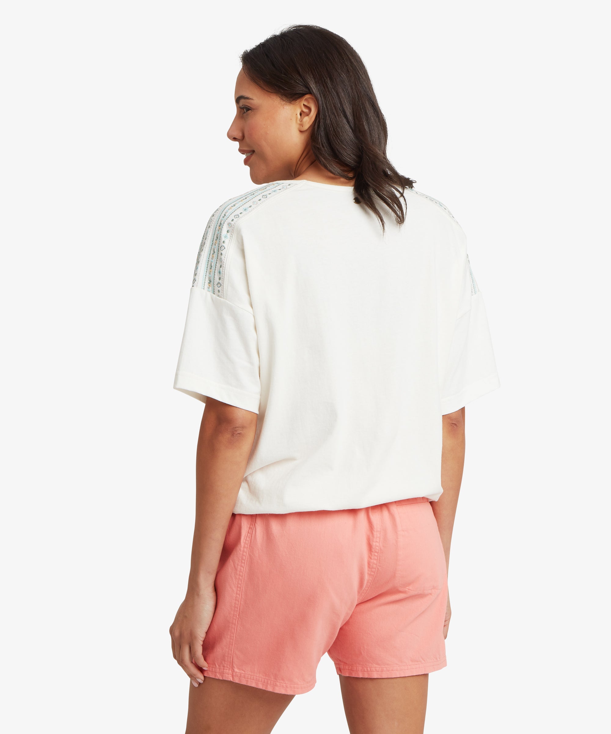 A back view of a person wearing a white short‑sleeved top with blue and white patterned stripes along the shoulders and sleeves, paired with loose‑fit coral shorts against a plain white background.