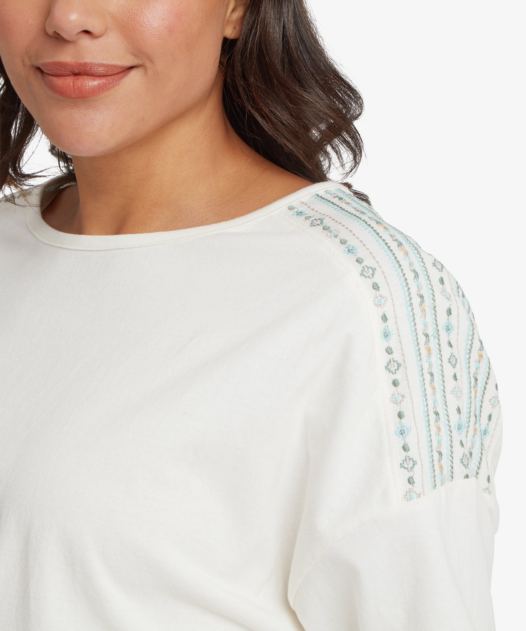 A close-up of a white top with a round neckline, featuring vertical embroidered detailing on the shoulder in blue and beige geometric patterns.