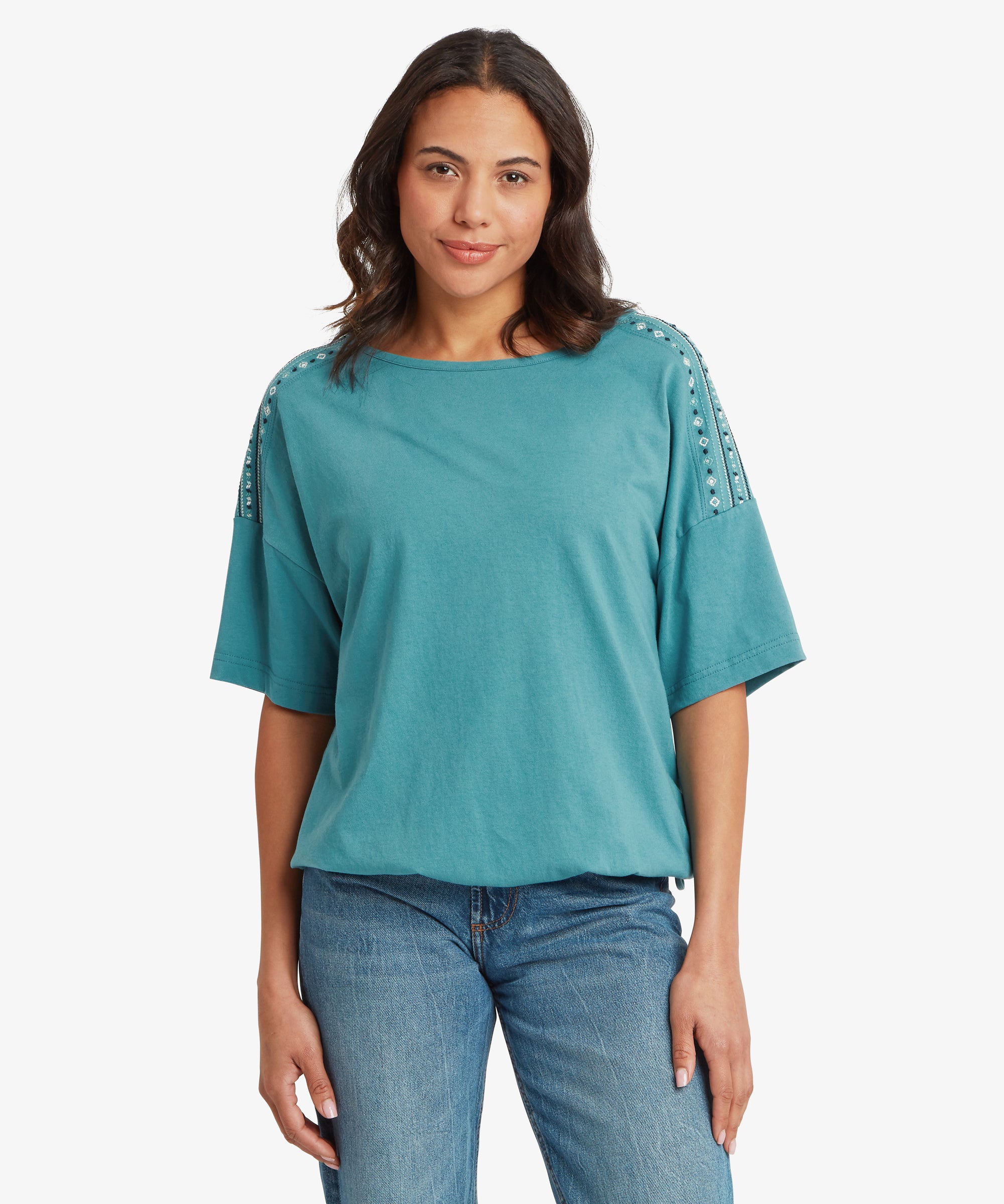 A back view of a person wearing a teal short‑sleeve top with white patterned stripes along the shoulder seams. The top drapes loosely over mid‑wash blue jeans, and their dark hair falls past the shoulders.