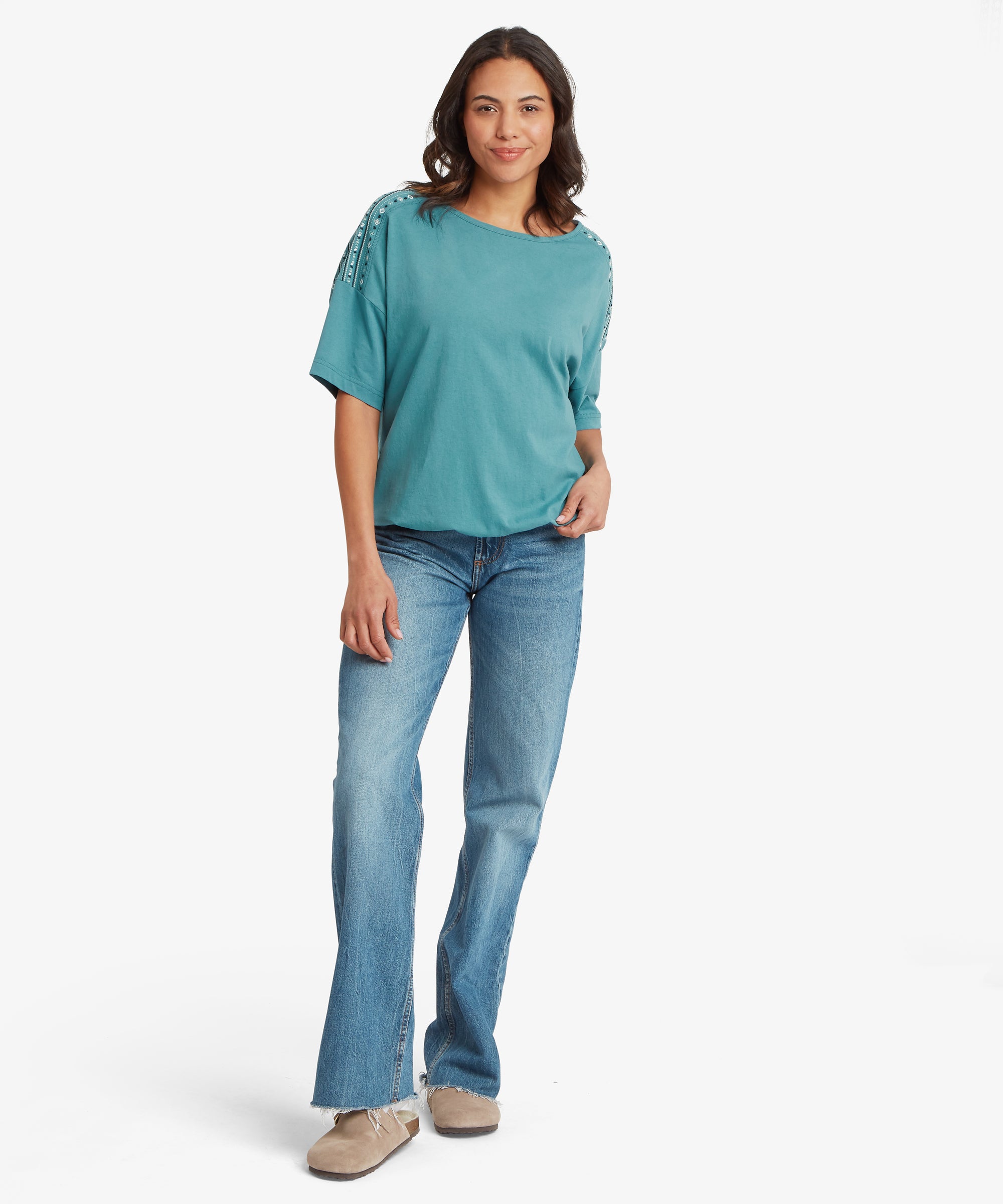 A person wearing a teal short‑sleeve top with a wide neckline and decorative metallic detailing along the shoulder seams, paired with straight‑leg blue jeans. The relaxed fit creates a casual everyday look.