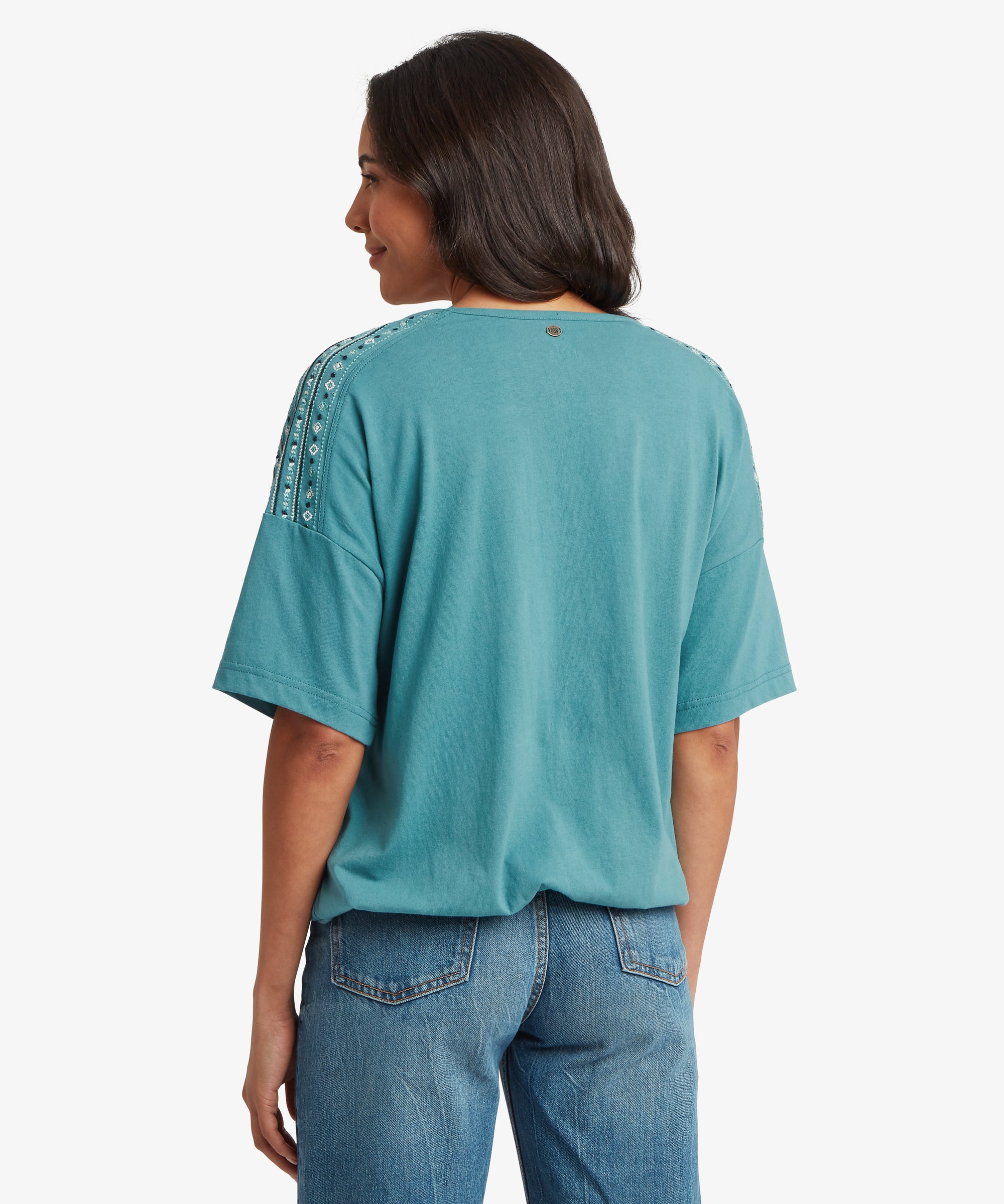 A person is shown from the back wearing a teal short‑sleeve top with decorative white patterned stripes running along the shoulder seams. The top has a relaxed fit and sits loosely over mid‑wash blue jeans with classic stitching and back pockets. Their dark hair falls past the shoulders, and the plain white background highlights the garment details.