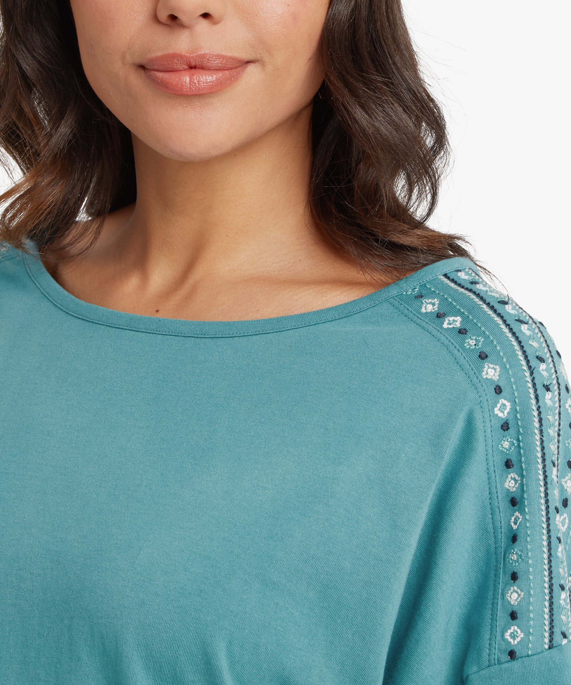 A close-up view of a person wearing a teal short‑sleeve top with a wide scoop neckline. The shoulders and sleeves feature intricate embroidered diamond motifs in white and dark blue, creating a textured, decorative panel. Only the lower half of the person’s face is visible, showing natural makeup and wavy dark hair framing the neckline.