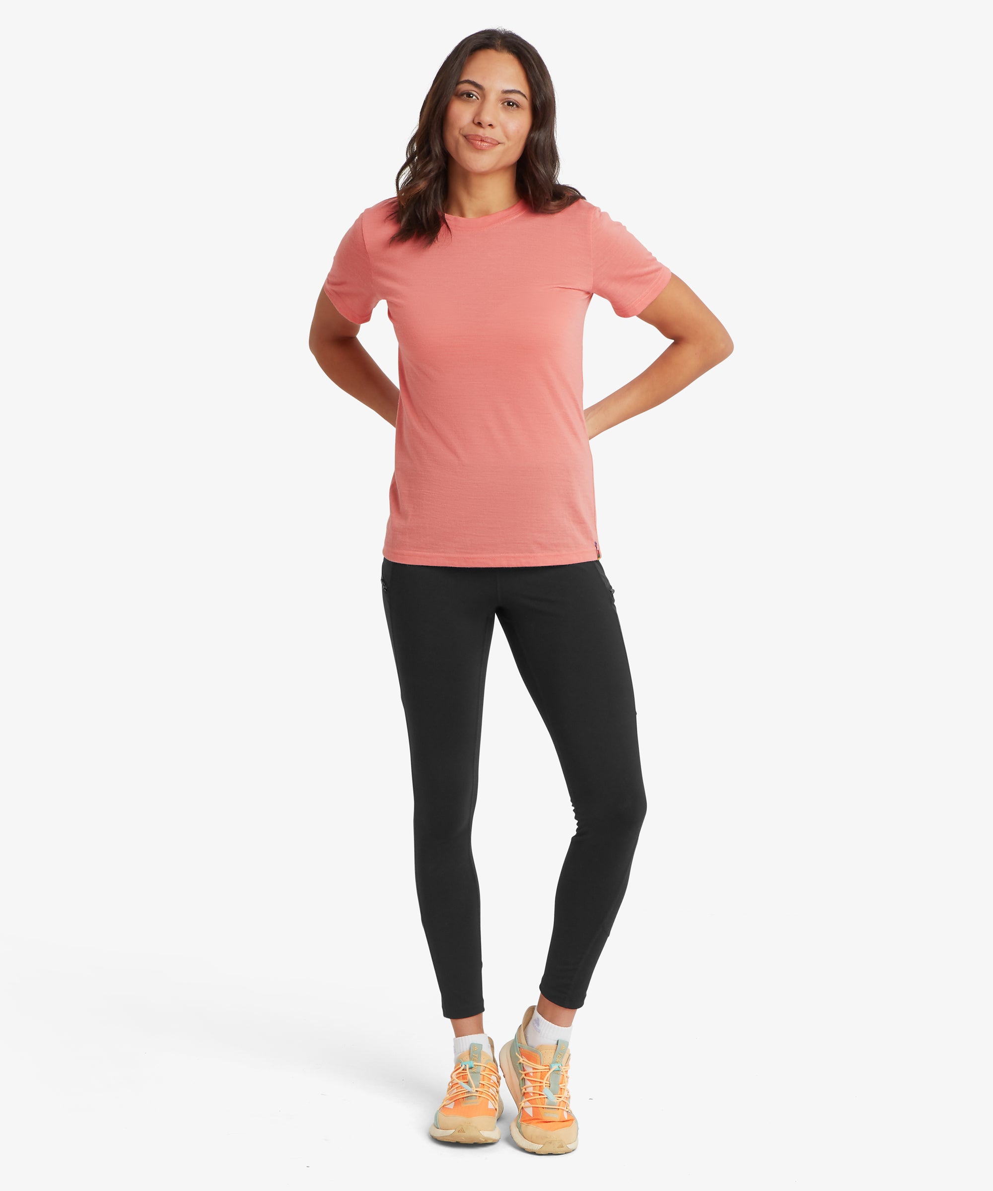 A person wearing a coral short‑sleeve T‑shirt, black leggings, and orange athletic shoes with white soles. They stand with hands behind their back against a plain white background, showing the full outfit and fit.