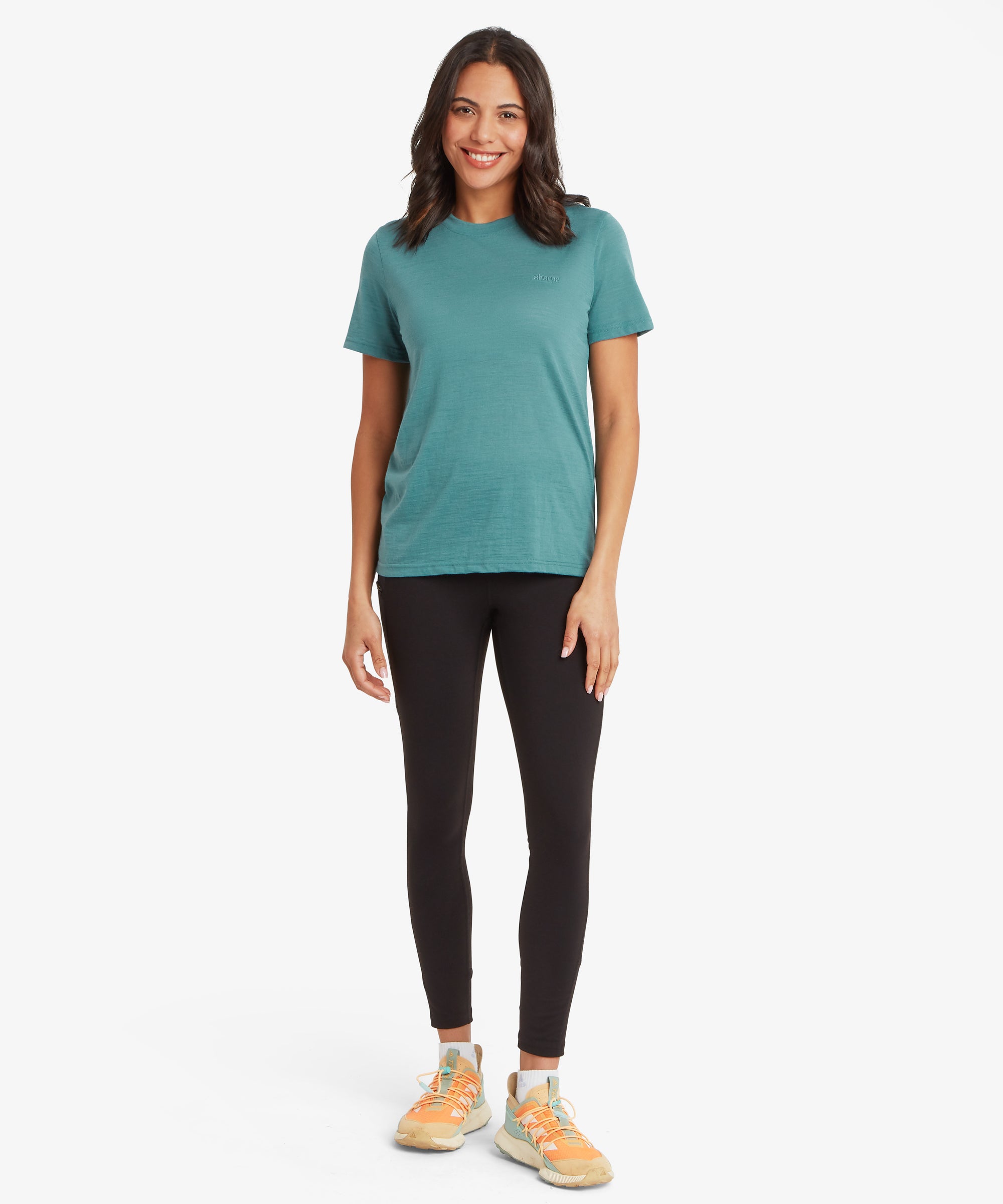 A person stands smiling against a plain white background wearing a teal short‑sleeve T‑shirt, black leggings, and orange athletic shoes with white soles and light blue laces. The relaxed fit and clean backdrop highlight the casual, sporty look.