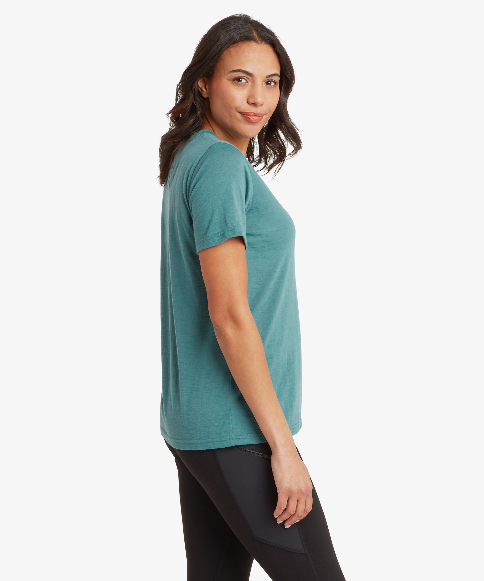 A person stands in a side pose wearing a teal short‑sleeve T‑shirt and black leggings with mixed‑texture panel detailing. The plain white background emphasises the fit and structure of both garments.