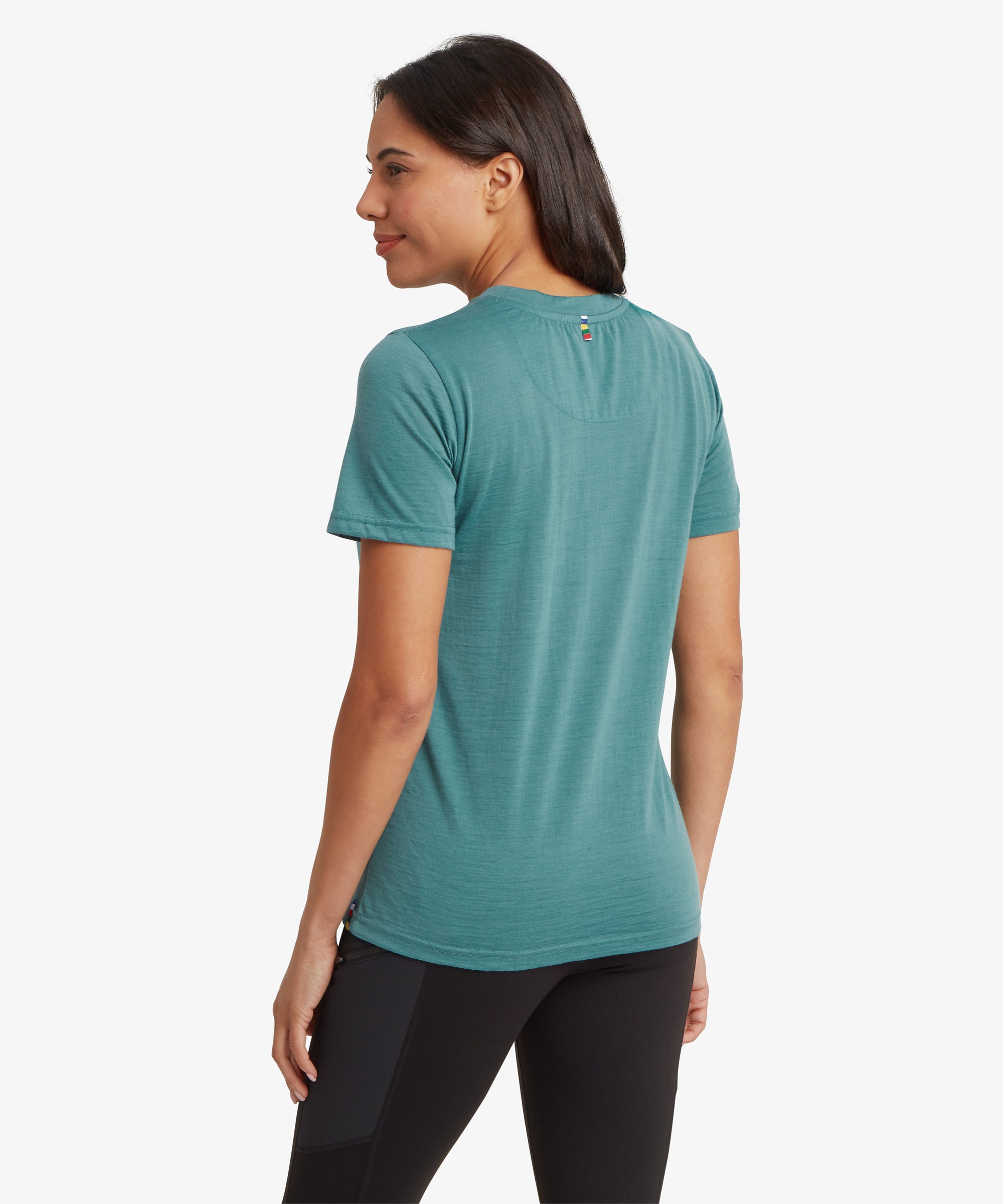 A back view of a person wearing a teal short‑sleeve T‑shirt featuring a small horizontal striped prayer flag detail below the neckline. They pair it with black leggings, and their long dark hair falls over the shoulders against a plain white background.