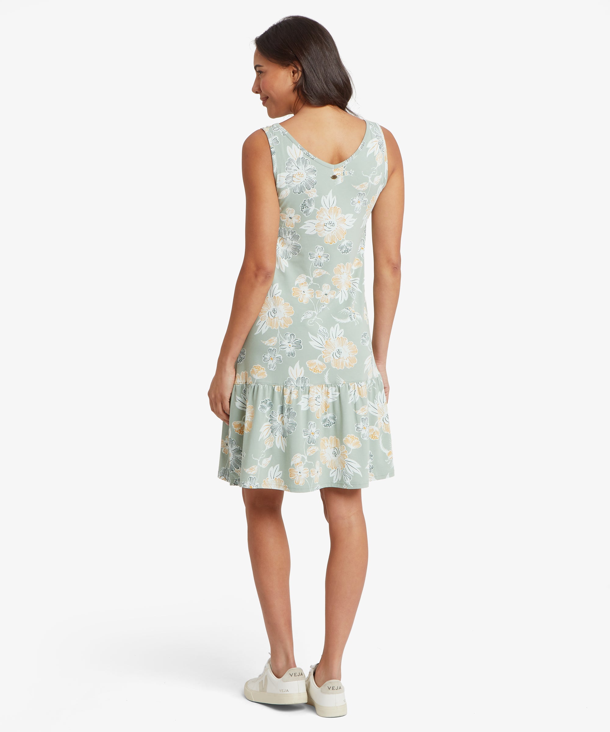 RŸckansicht eines grŸnen, Šrmellosen Blumenkleids mit V-fšrmigem RŸckenausschnitt und gerŸschtem Saum, kombiniert mit wei§en Sneakers.