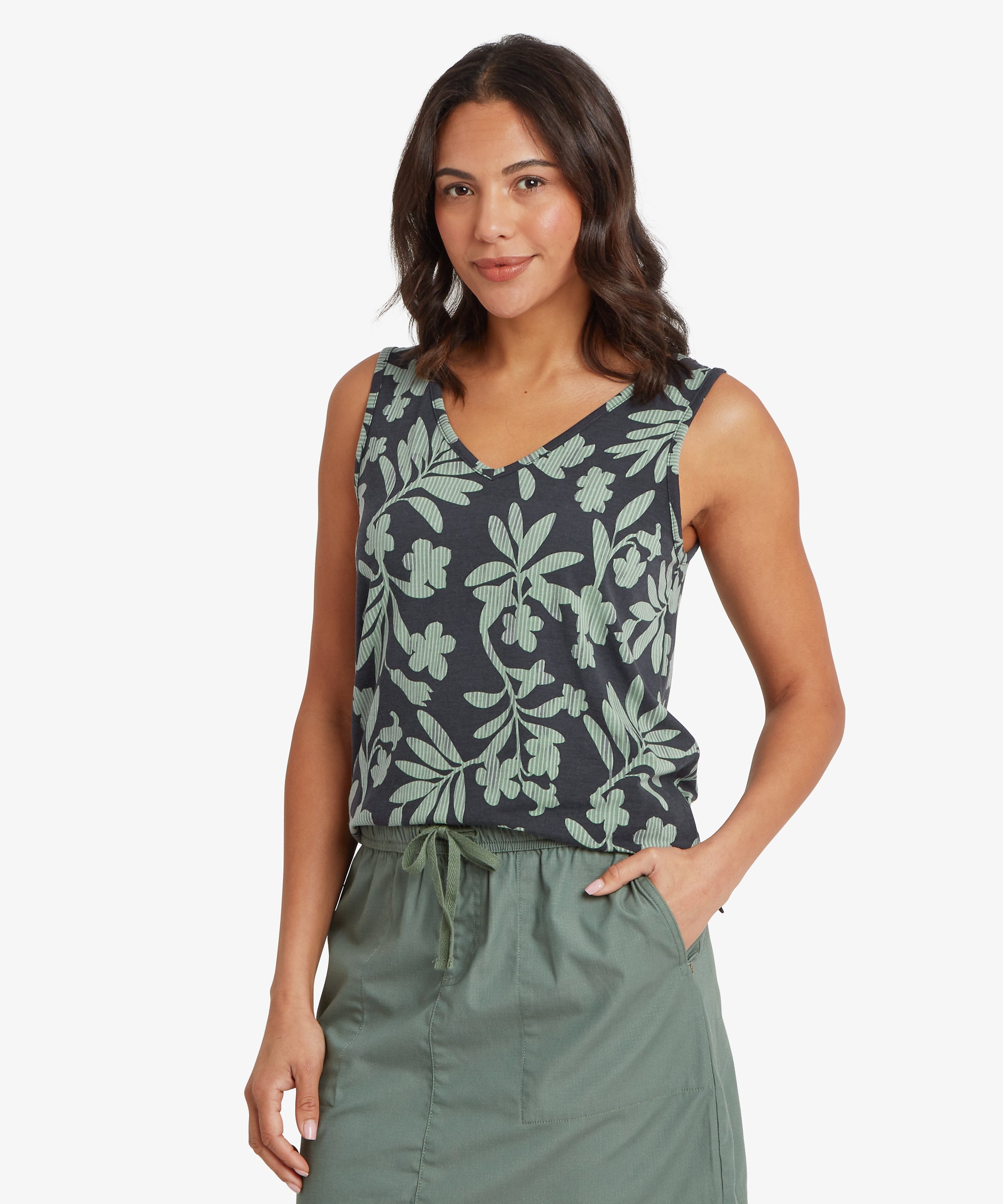 Woman facing forward wearing a sleeveless green V-neck top with a relaxed fit through the body, paired with a green drawstring skirt. One hand rests in her skirt pocket and she has a soft, neutral smile.
