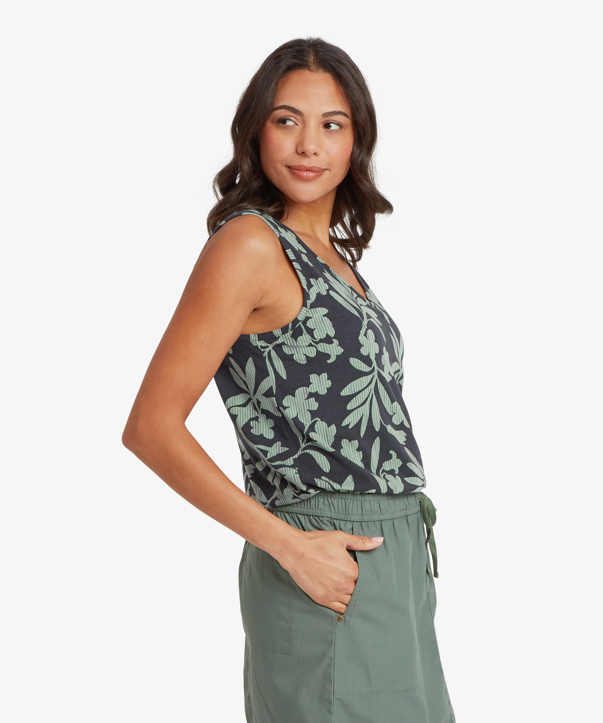 Woman shown from a three-quarter side angle wearing a sleeveless green V-neck top tucked lightly into a green skirt. One hand is placed in her pocket, highlighting the armhole shape and relaxed fit across the torso.