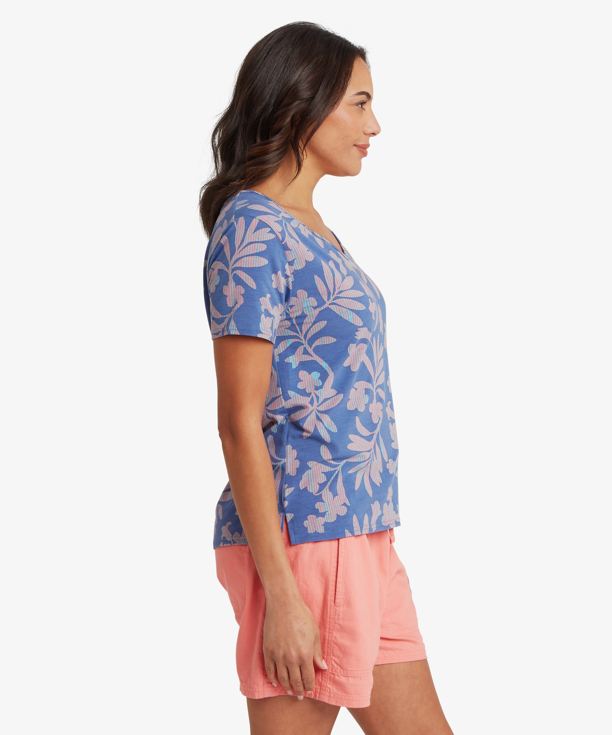 Side view of a woman wearing a blue floral V-neck T-shirt and coral shorts, showing the straight silhouette, lightweight fabric, and short sleeves that fall to mid-upper arm; her gaze is forward and posture relaxed.