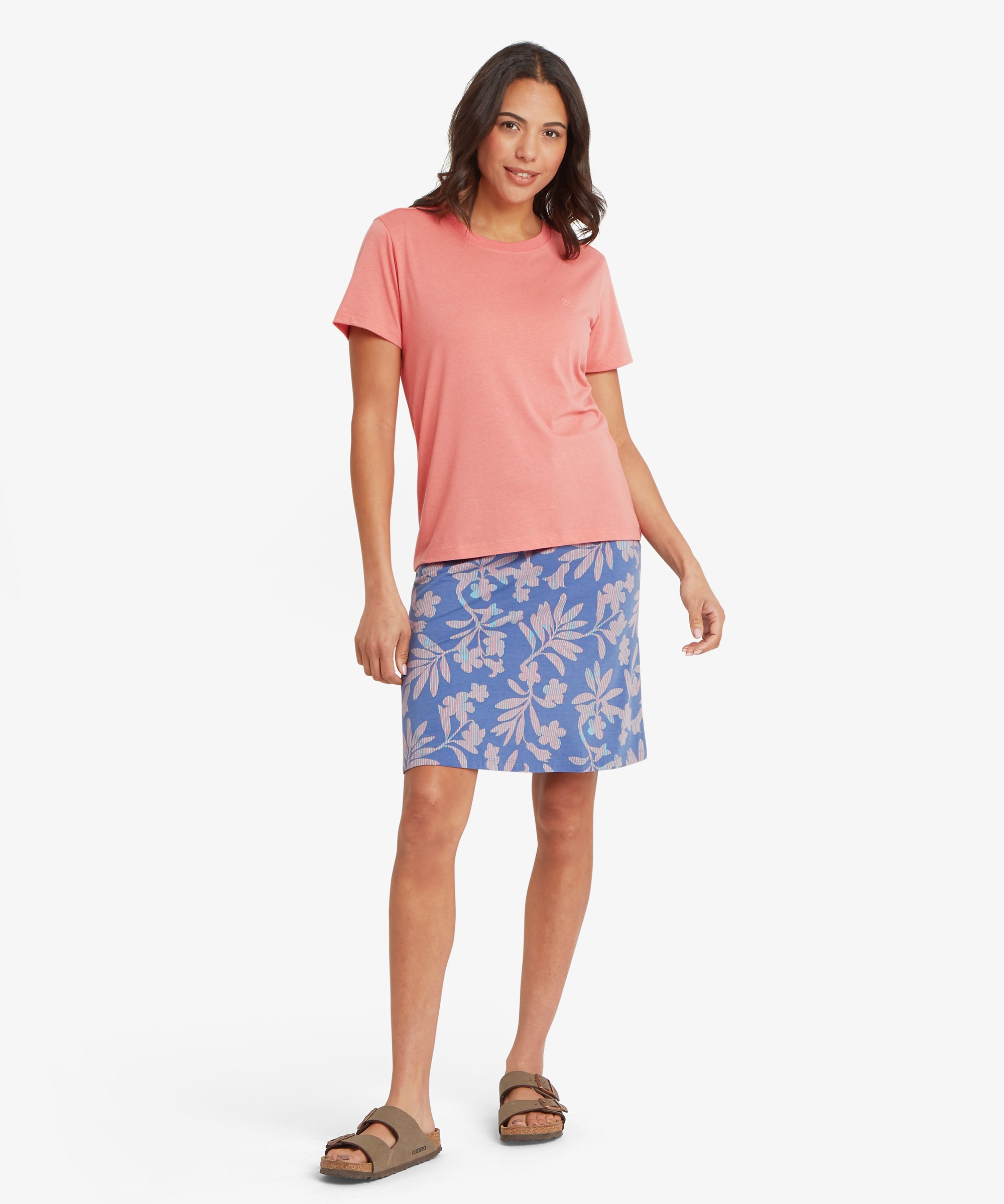 Ganzkšrperaufnahme einer Frau, die einen blauen A-Linien-Rock mit Blumenmuster, knielangem Saum und lockerer Passform trŠgt, kombiniert mit einem korallenfarbenen Kurzarm-T-Shirt und flachen Sandalen.