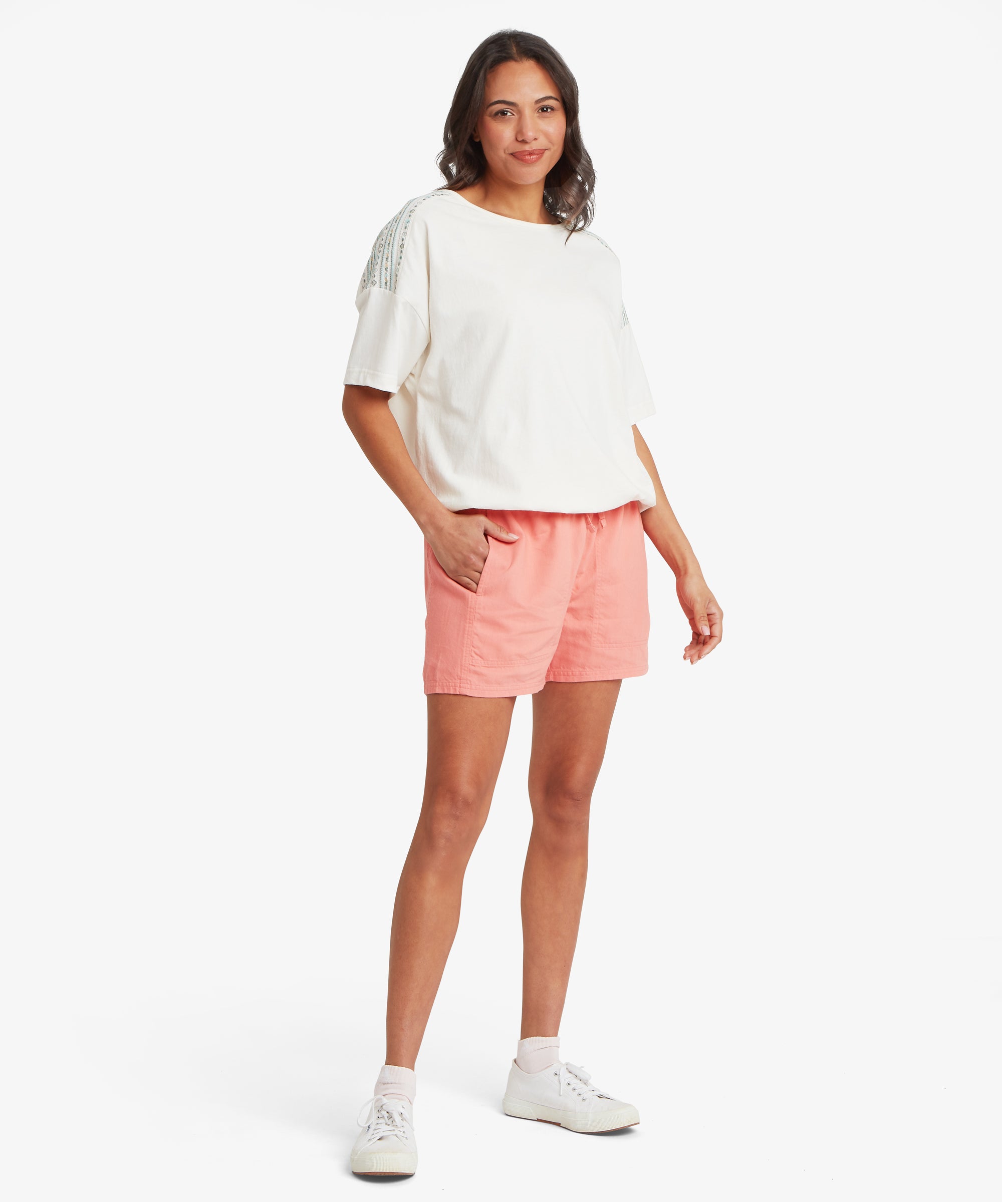 Full-body view of a woman wearing coral-pink elastic-waist shorts with a loose fit through the hips and thighs, styled with a white short-sleeve T-shirt and trainers.