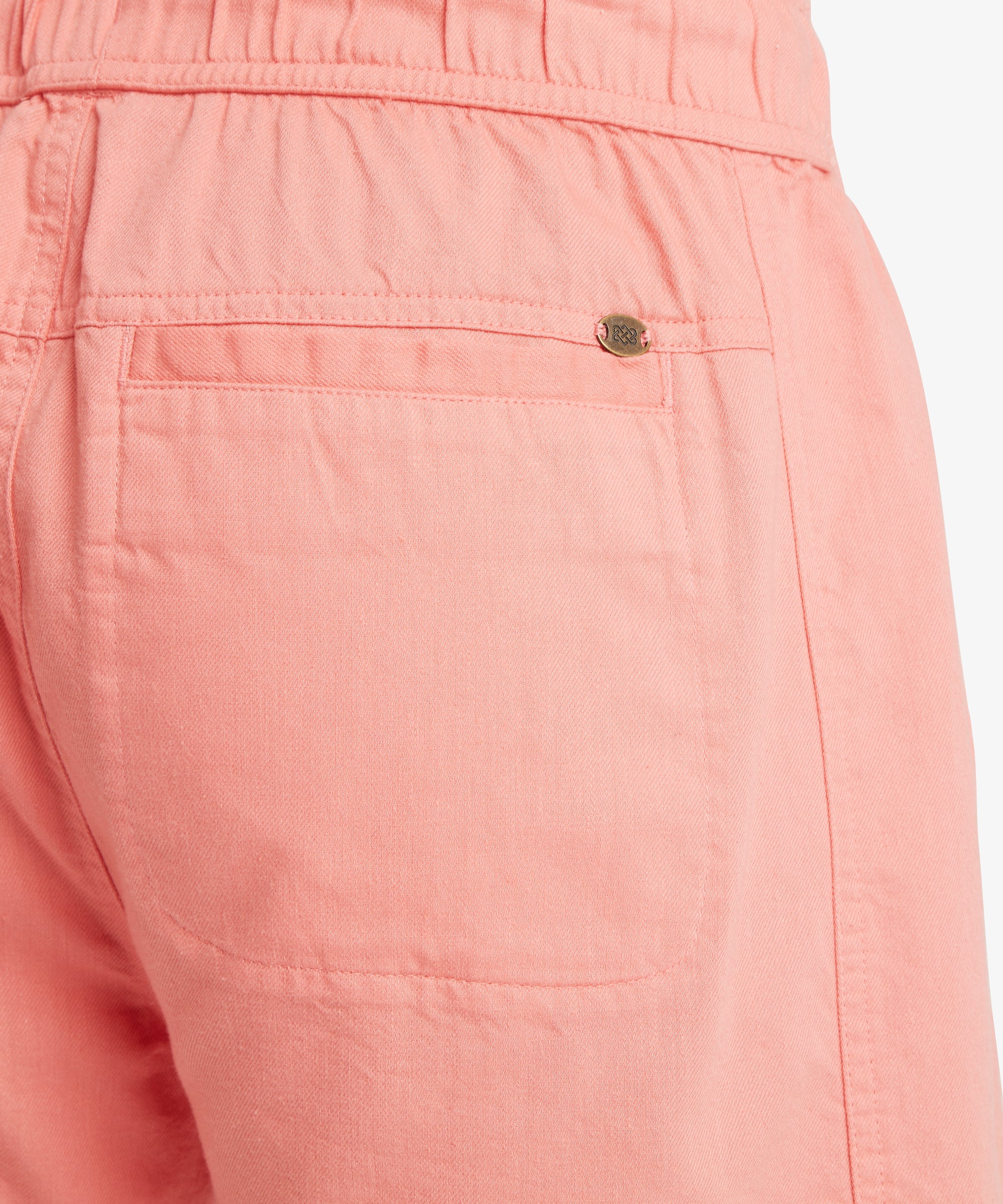 Close-up of the back of coral-pink shorts showing a single welt-style back pocket with a small, metal endless knot logo tab.