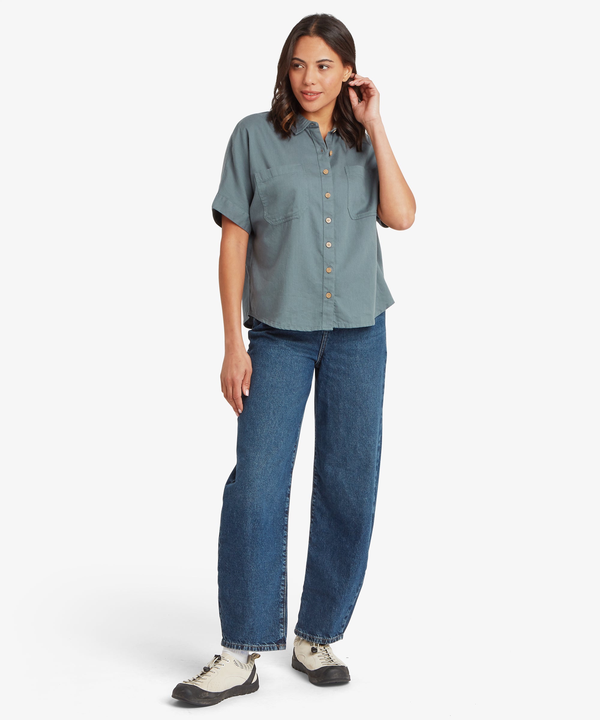 Full-body view of a woman wearing a relaxed-fit blue-grey short-sleeve shirt with a loose silhouette, paired with straight-leg jeans and trainers.