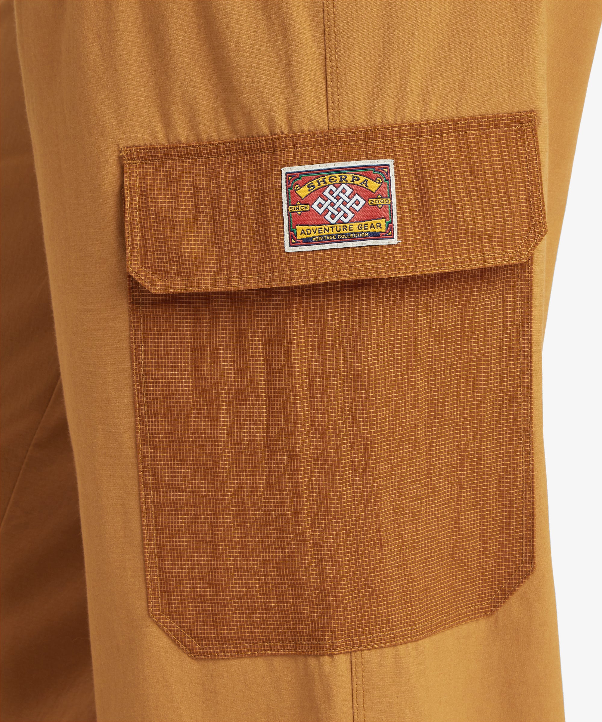A close-up of a textured brown‑orange pocket with a grid‑patterned fabric and a stitched flap. The pocket features a colourful embroidered patch with branding and a traditional knot motif, highlighting the garment’s outdoor‑inspired detailing.
