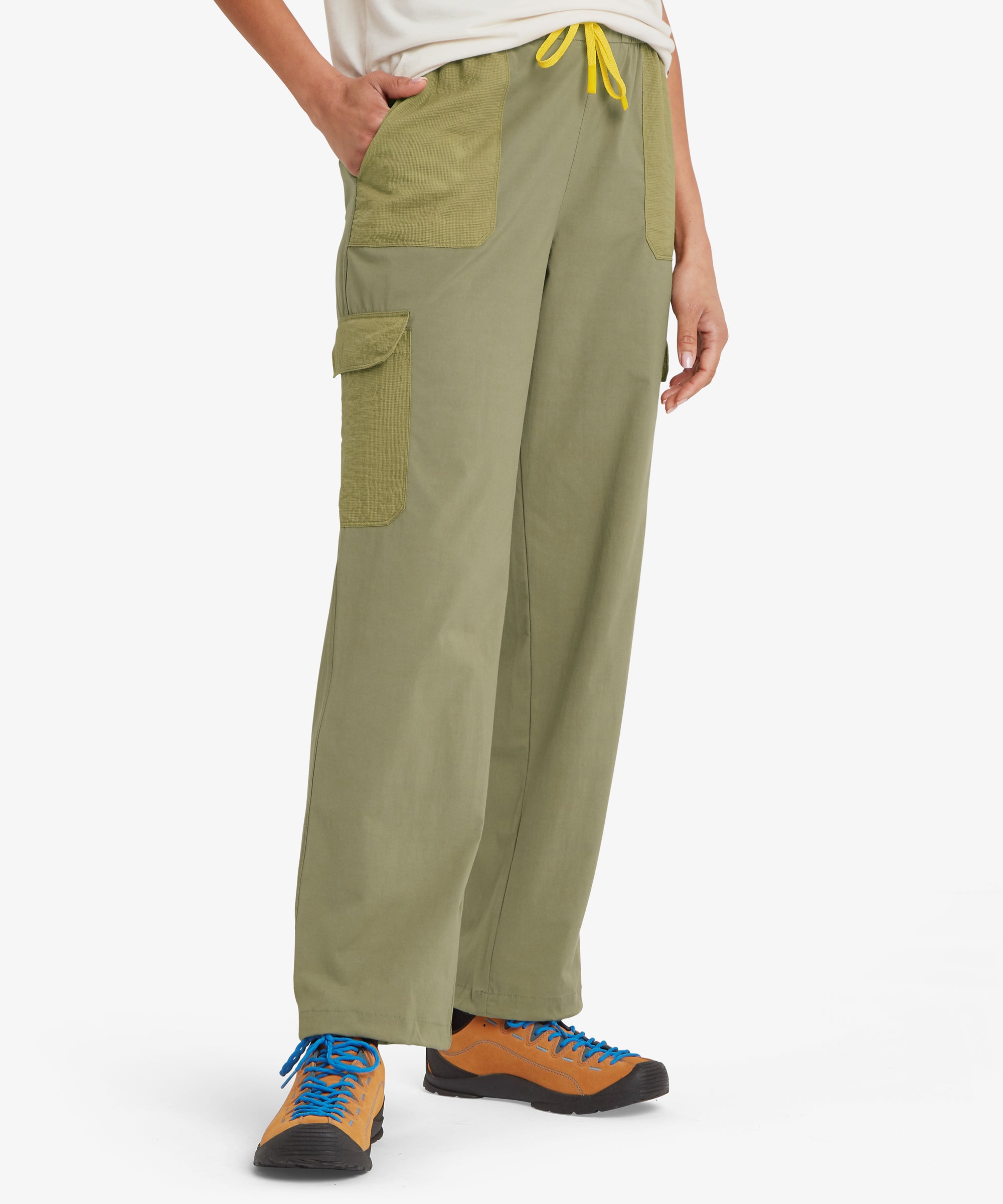 A person facing forward wearing a plain white short‑sleeve T‑shirt and olive cargo trousers with contrasting green pockets and a yellow drawstring. Tan hiking shoes with blue laces complete the outfit, shown against a plain white background.