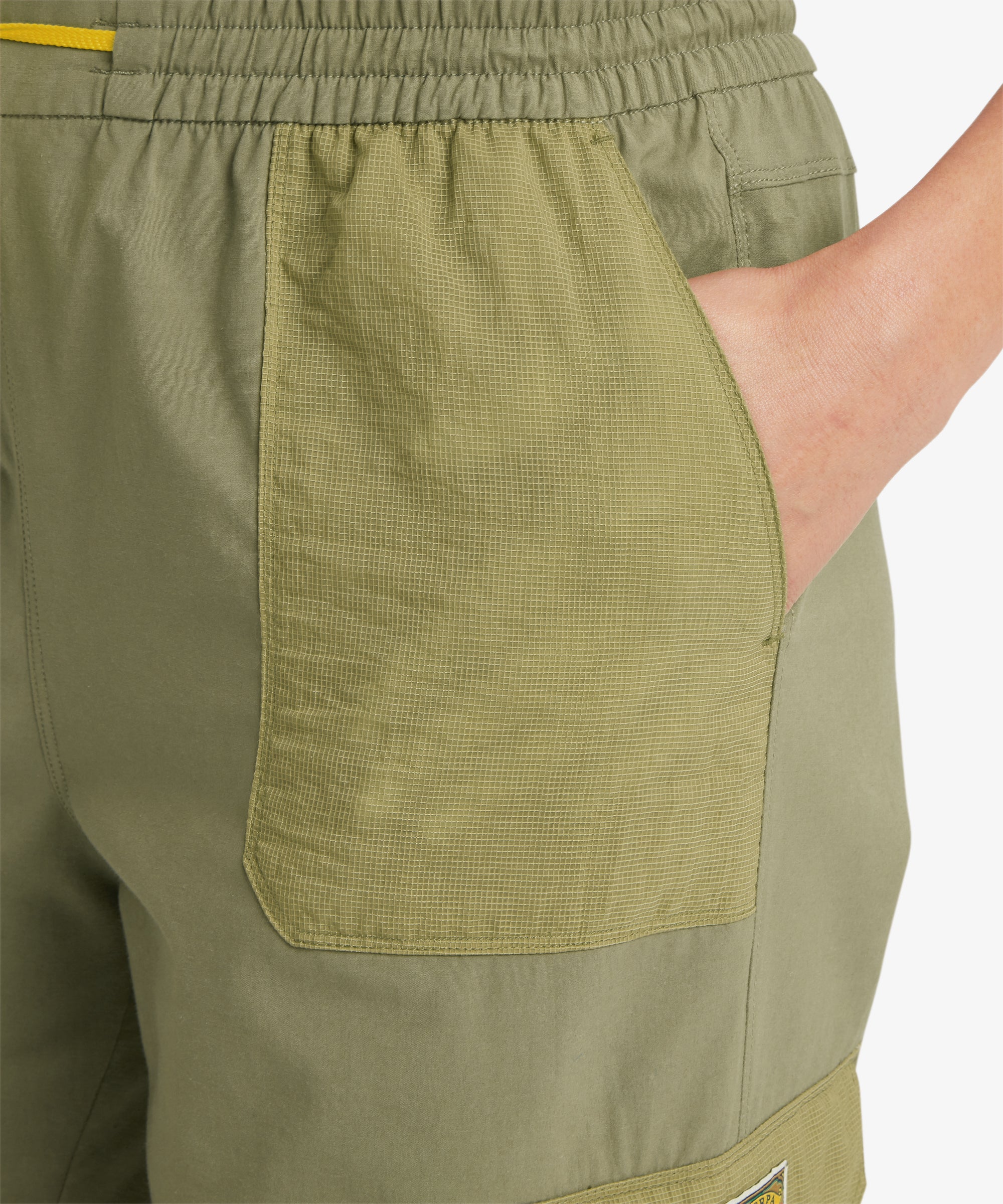 A person wearing olive cargo trousers with large front and side patch pockets, including a flap pocket on the thigh. The trousers are paired with a white top and orange hiking shoes, highlighting the functional outdoor style.