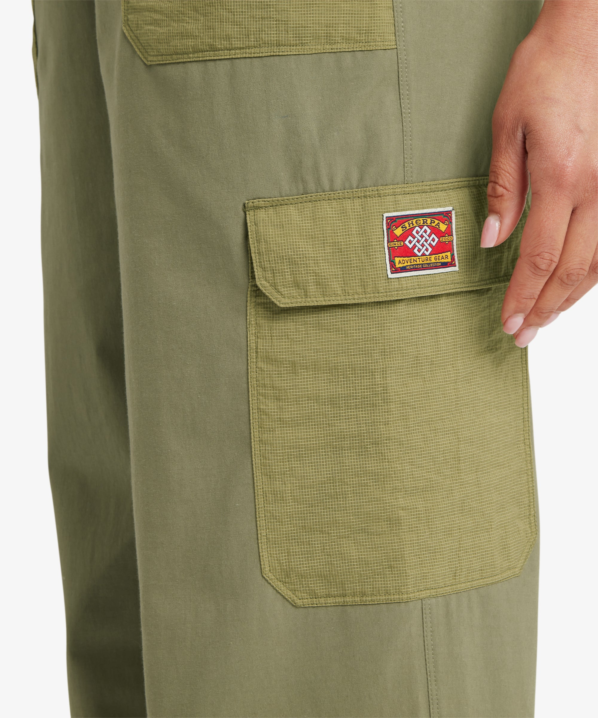 A close-up of green shorts with an elastic waistband and a contrasting textured patch pocket in a grid‑patterned fabric. A hand rests inside the pocket, showing the construction and material contrast.