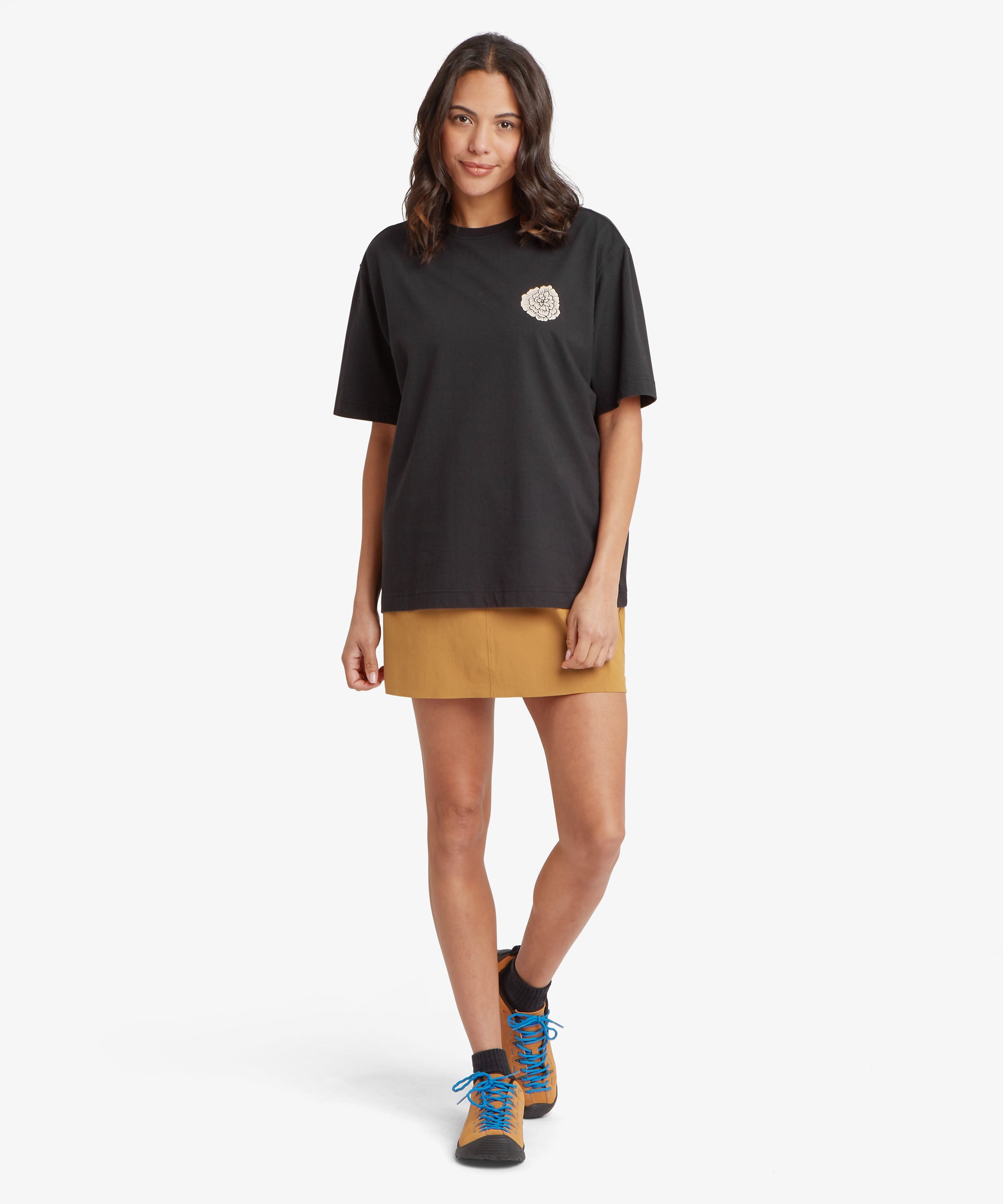 A person wearing a black oversized T‑shirt with a small circular chest graphic, a mustard skirt, and tan hiking boots with blue laces. The plain white background highlights the casual, outdoors‑inspired look.