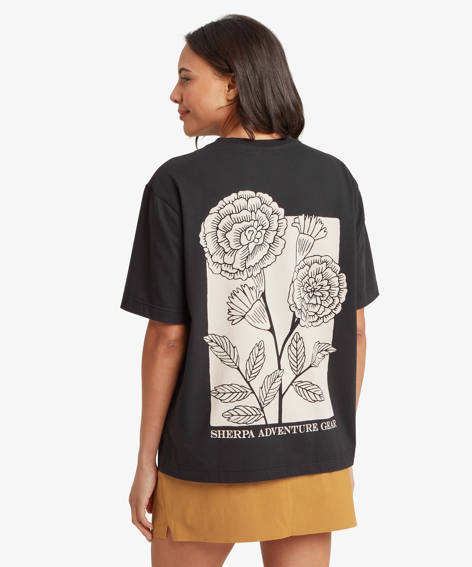 A person wearing a black T‑shirt with a large floral line‑art design on the back and a small floral emblem on the front. The back print features detailed flowers and leaves with branding beneath, paired with a mustard skirt.